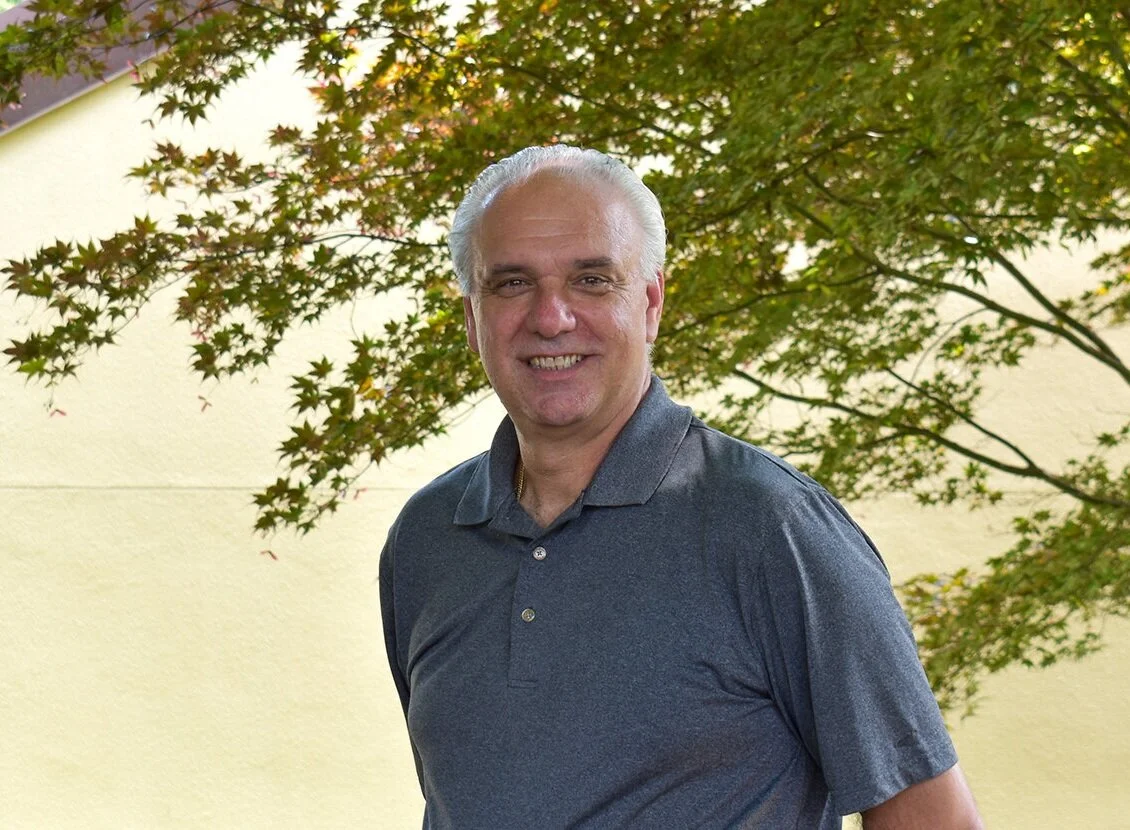 Smiling man with white hair in a dark gray polo shirt standing outdoors in front of a tree with green leaves.