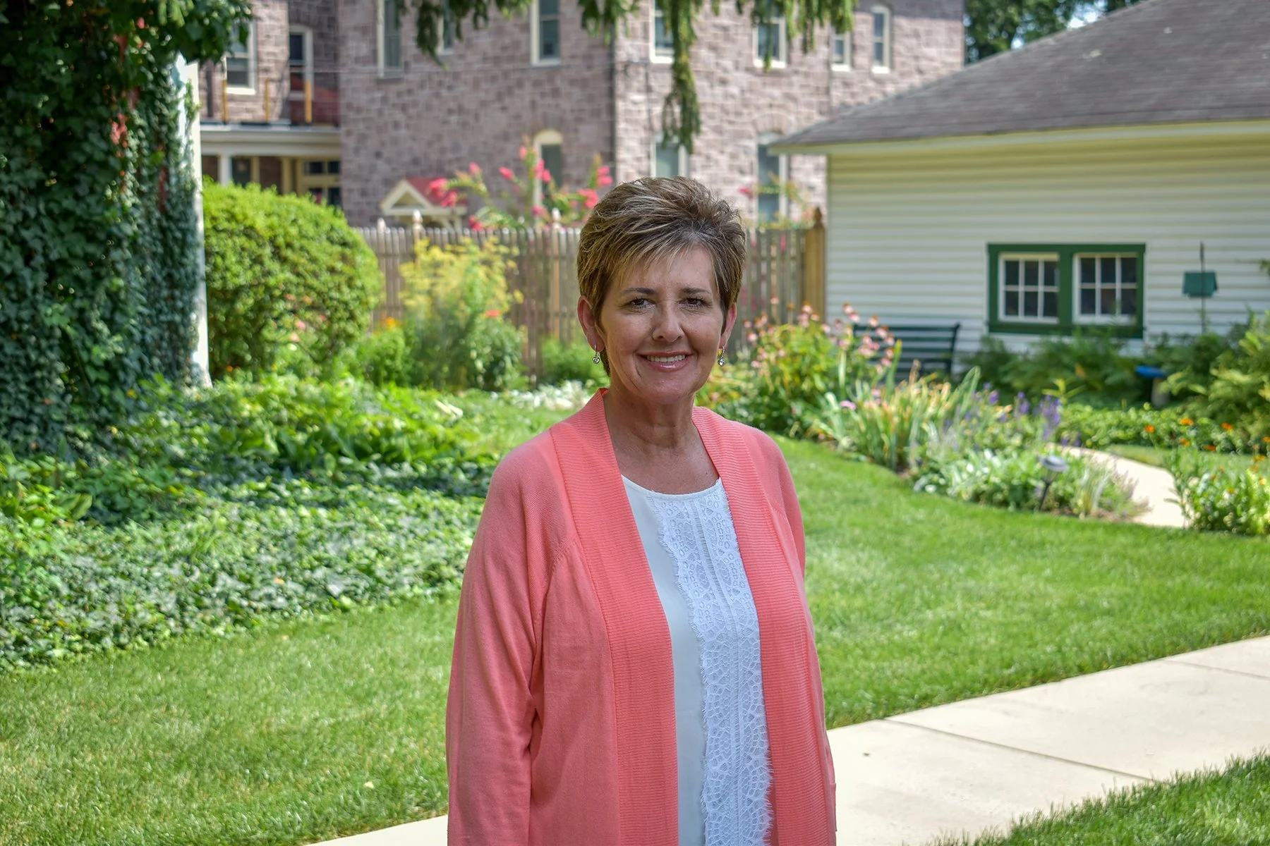 Smiling woman with short brown hair wearing a pink cardigan and white top standing in a lush green garden with flowering plants, surrounded by a residential neighborhood.