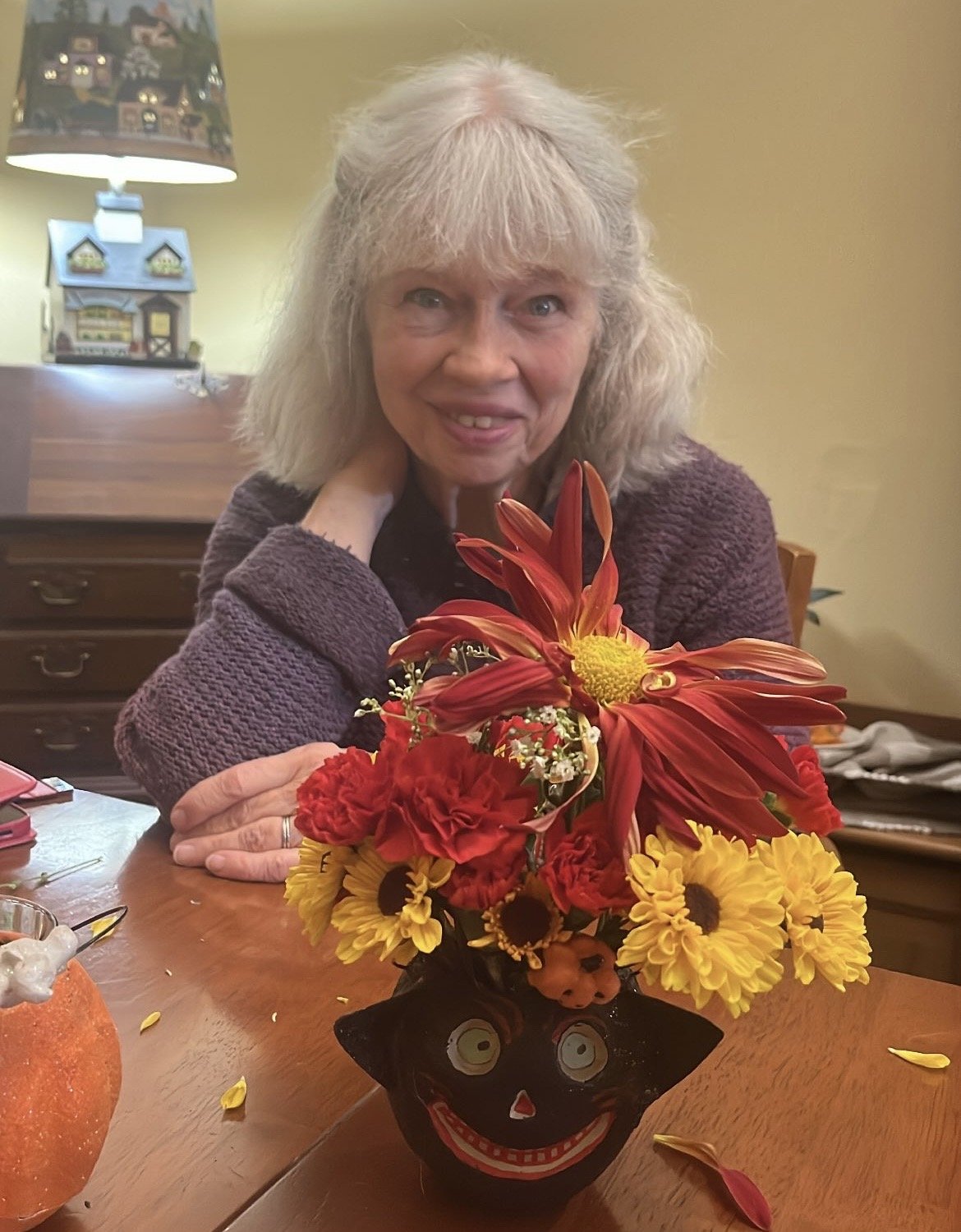 A woman with blonde hair sitting at a table with a colorful fall flower arrangement in a black vase with a smiling face design.