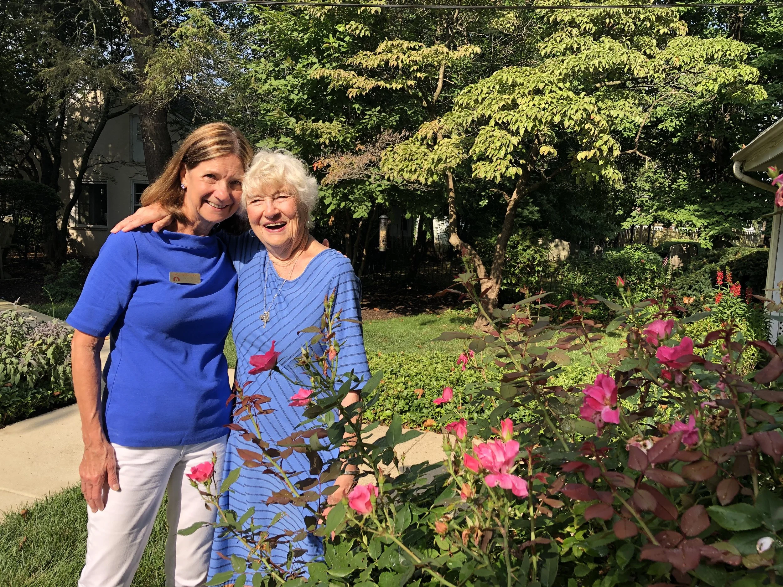 Two women smiling and hugging outdoors in a garden with blooming pink flowers and trees in the background, sunlight shining.