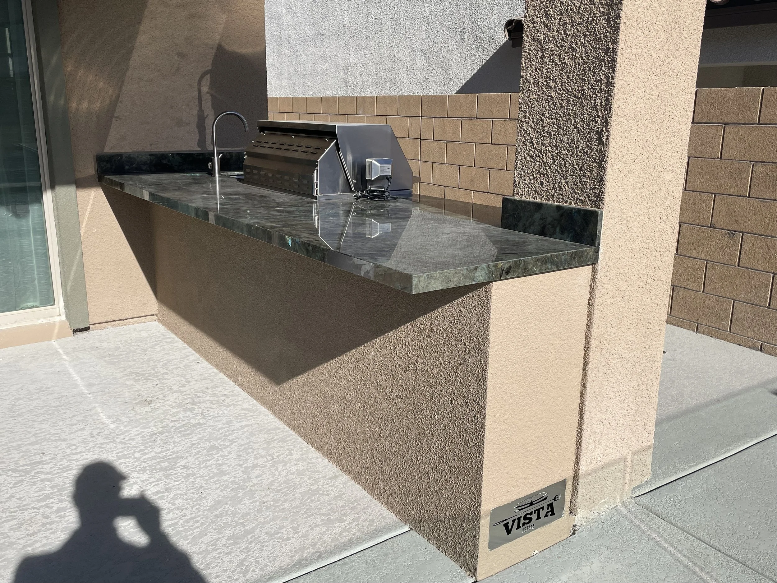 Outdoor kitchen counter with built-in stainless steel grill and faucet, situated on a concrete patio under bright sunlight.