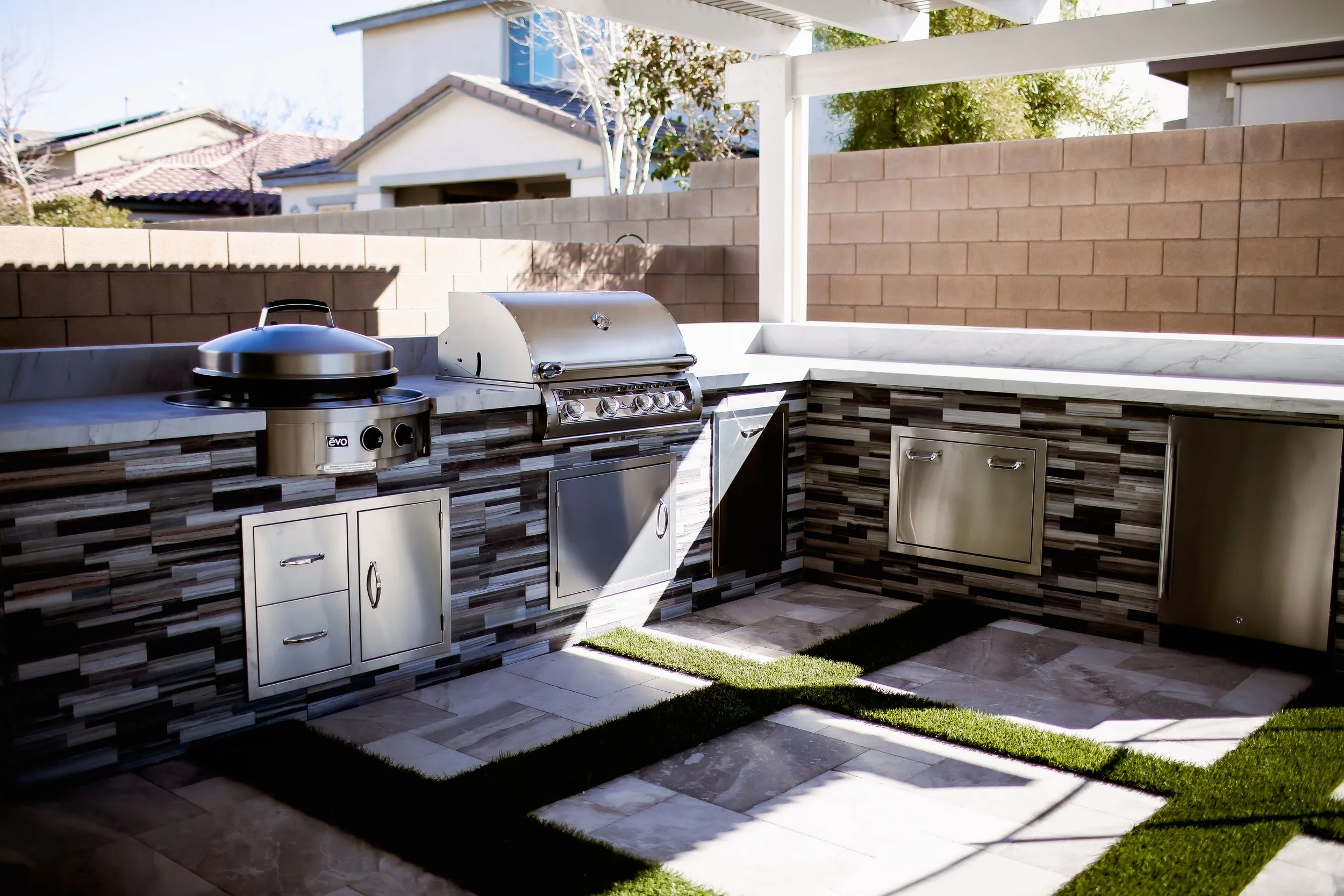 Outdoor kitchen with grill and appliances, stone countertops, modern design, brick wall, sunlight casting shadows on tiled floor and artificial grass accents.