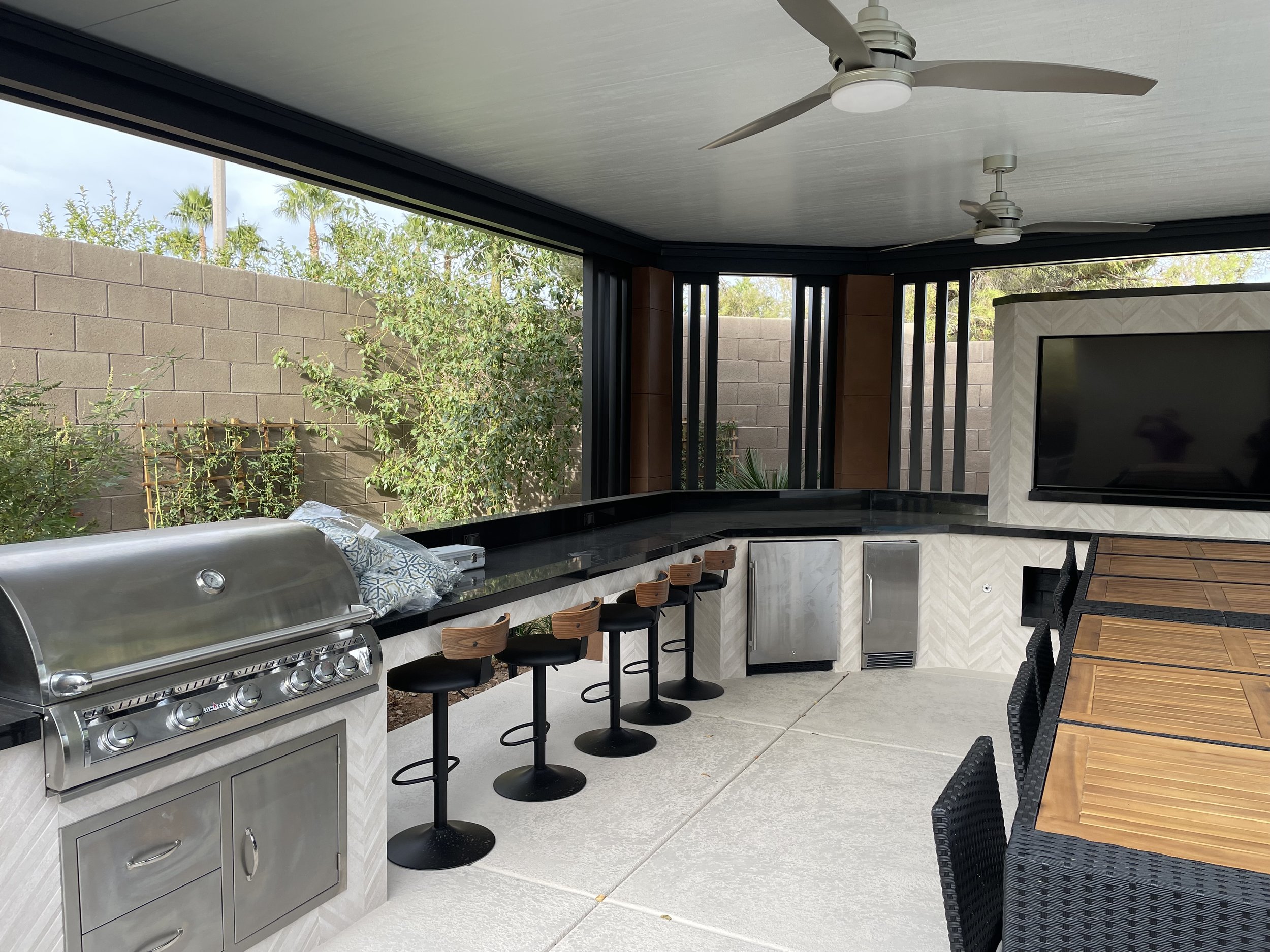 Outdoor kitchen with stainless steel grill, bar stools, TV, ceiling fans, and patio furniture, surrounded by plants and wooden fencing.