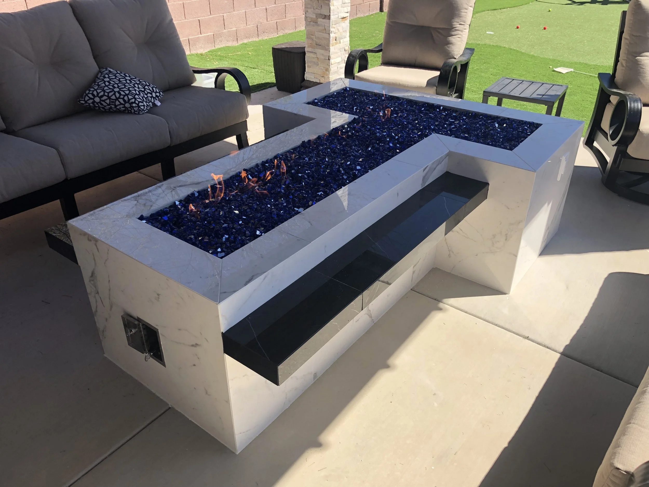 Outdoor L-shaped fire pit with blue glass rocks, surrounded by beige cushioned patio furniture on a concrete patio.
