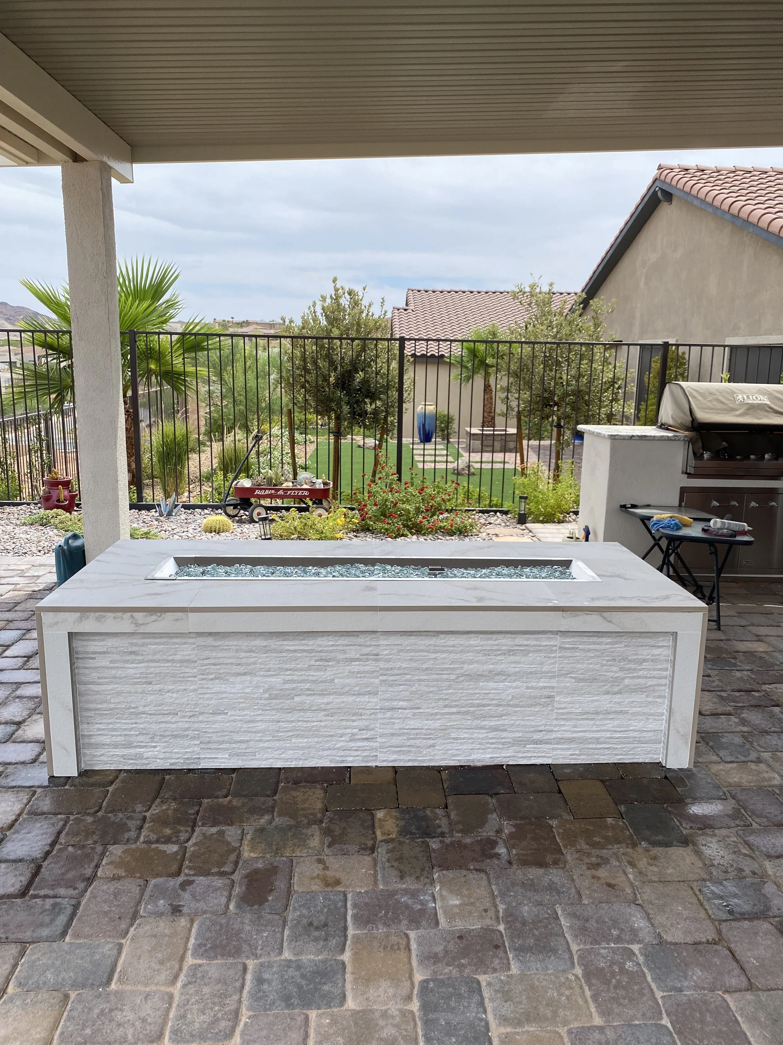 Outdoor patio area with a stone fire pit, patio cover, and paved flooring. In the background, there's a fenced yard with palm trees, shrubs, and a small wagon, with neighboring houses visible.