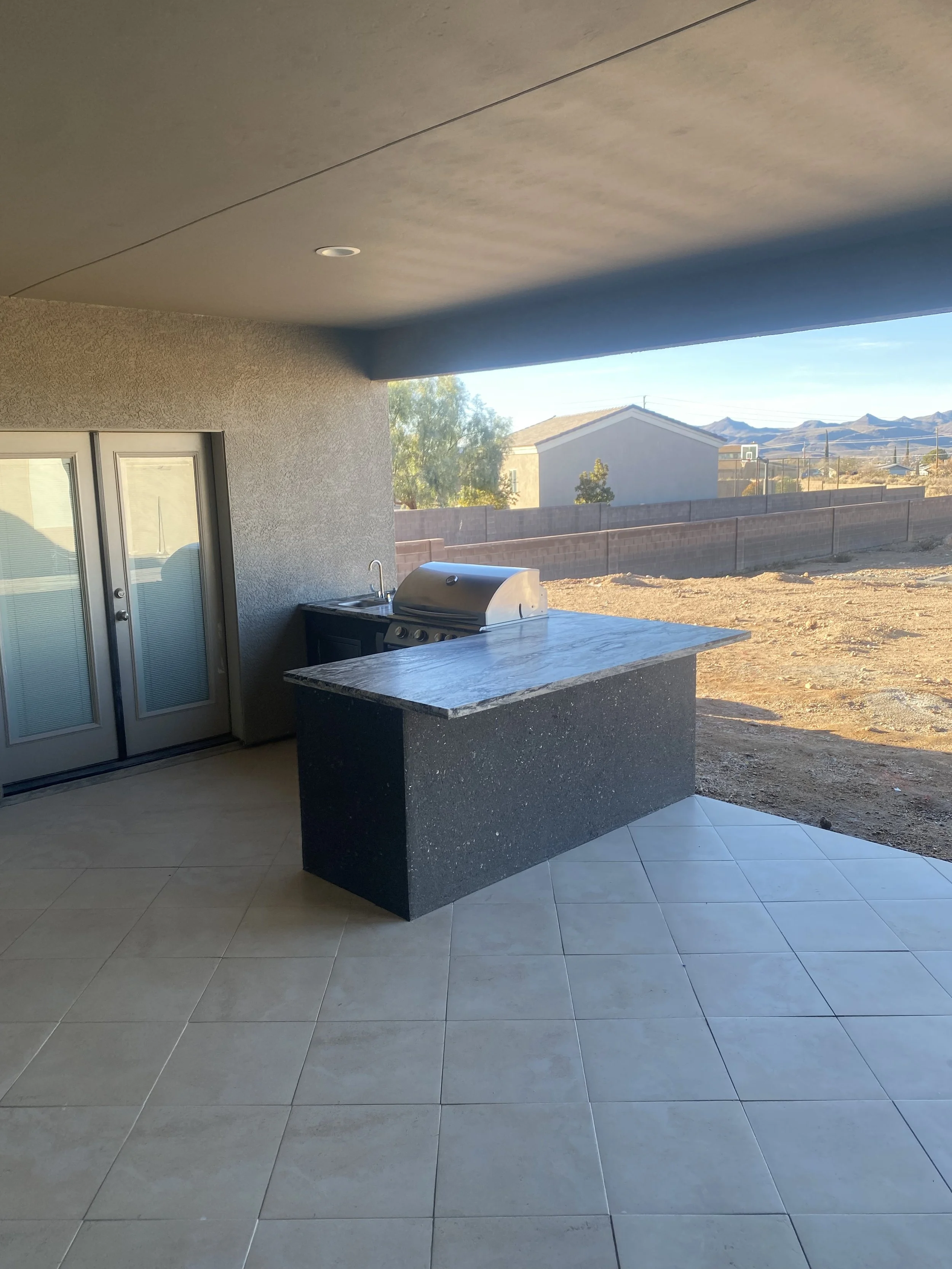 Covered patio with outdoor kitchen, featuring a grill and countertop, overlooking a walled backyard and distant mountains.