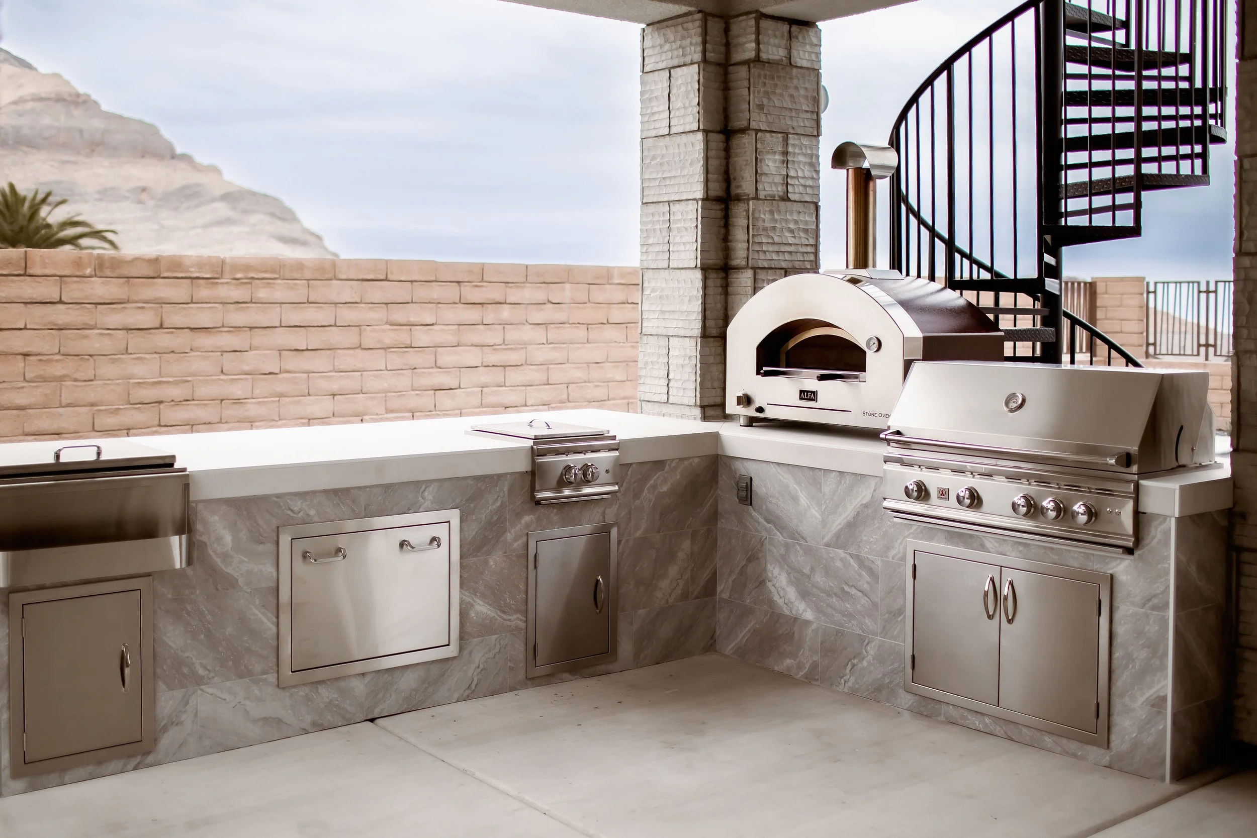 Outdoor kitchen setup with stainless steel appliances, including a built-in grill, oven, and storage cabinets, next to a spiral staircase and against a brick wall backdrop.