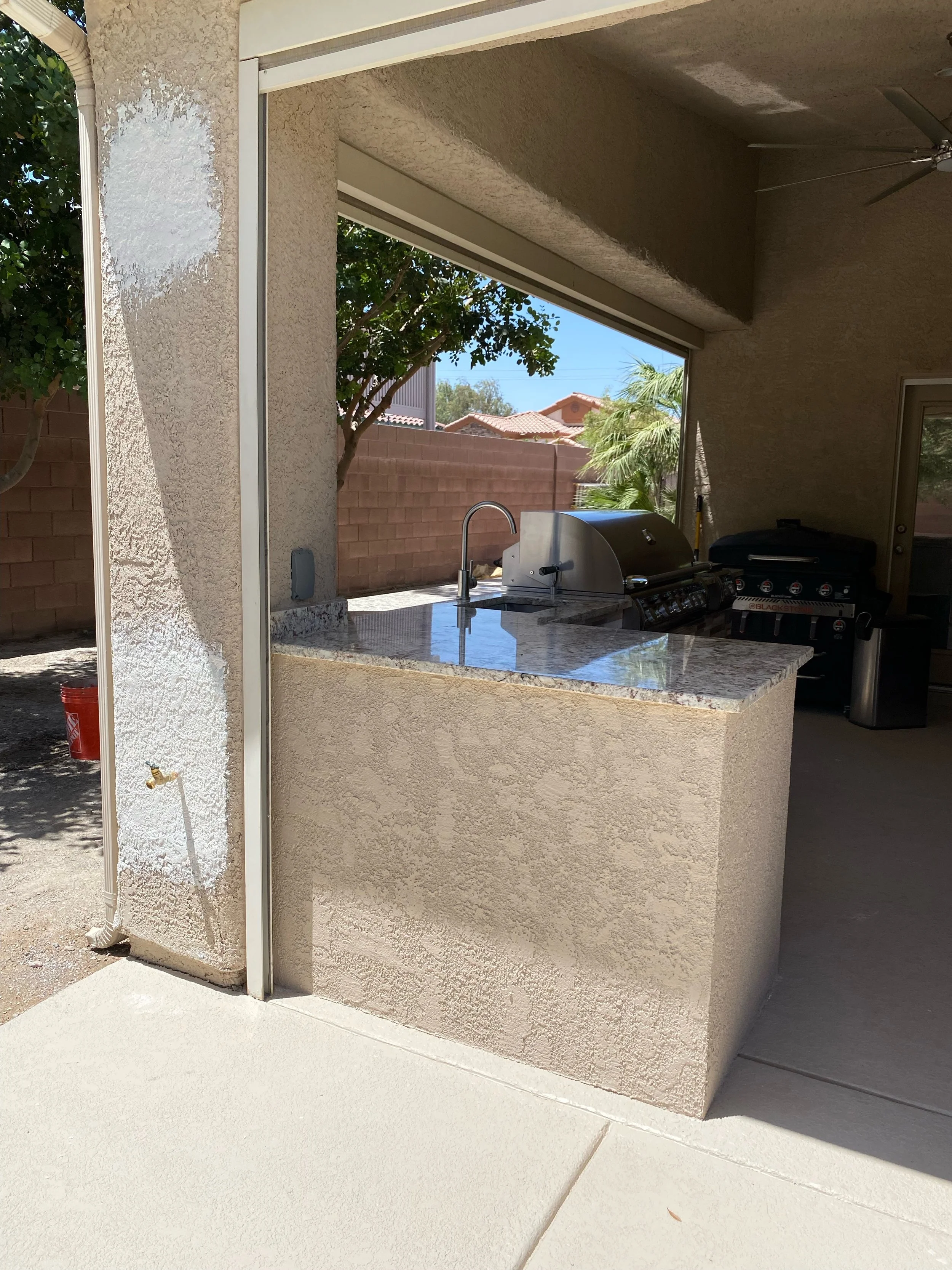 Outdoor covered patio area with a built-in grill and countertop, featuring stucco walls, ceiling fan, and partial view of the backyard with trees and a brick wall.