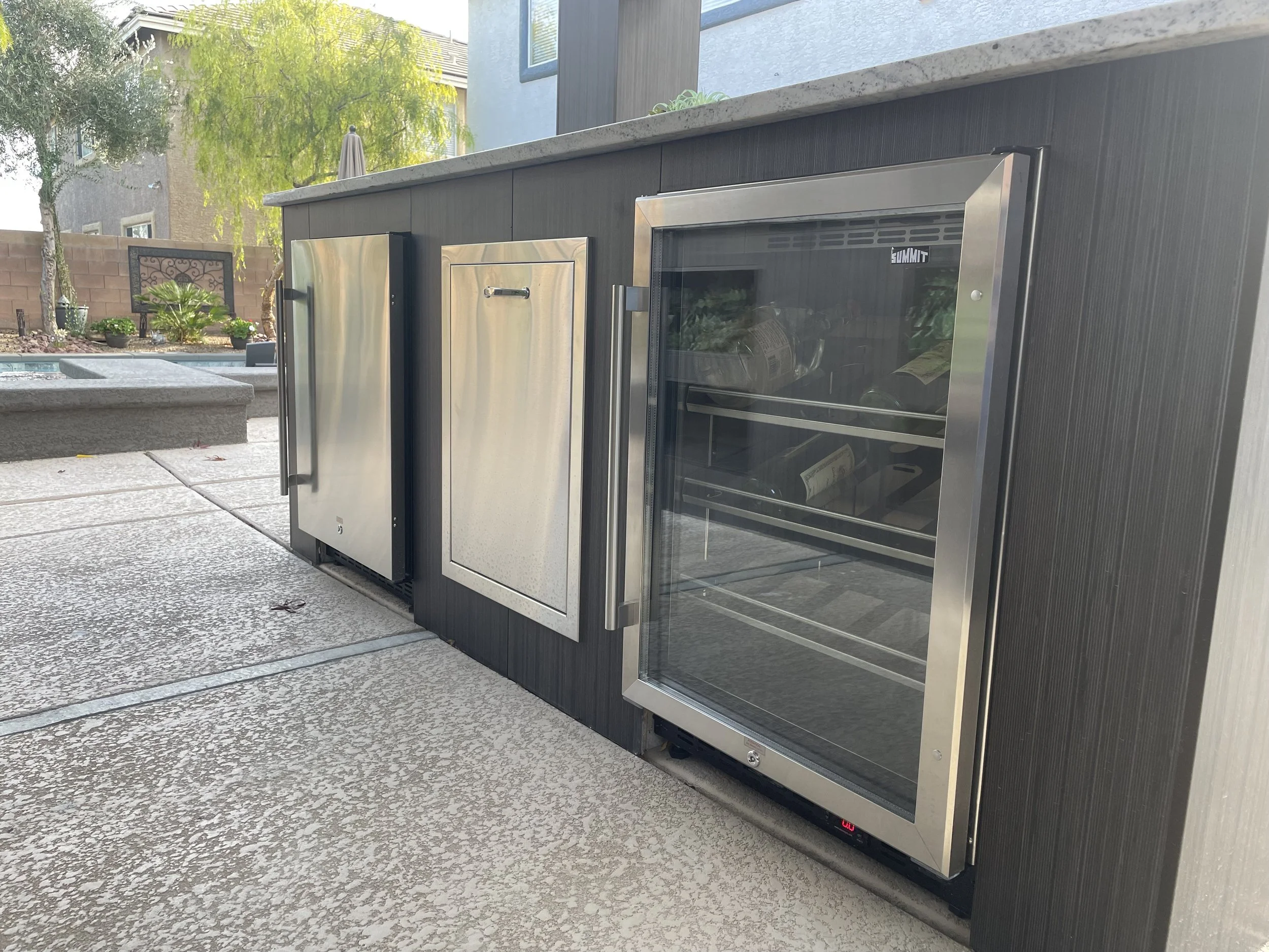 Outdoor kitchen with stainless steel mini fridge, cabinet, and wine cooler on patio.