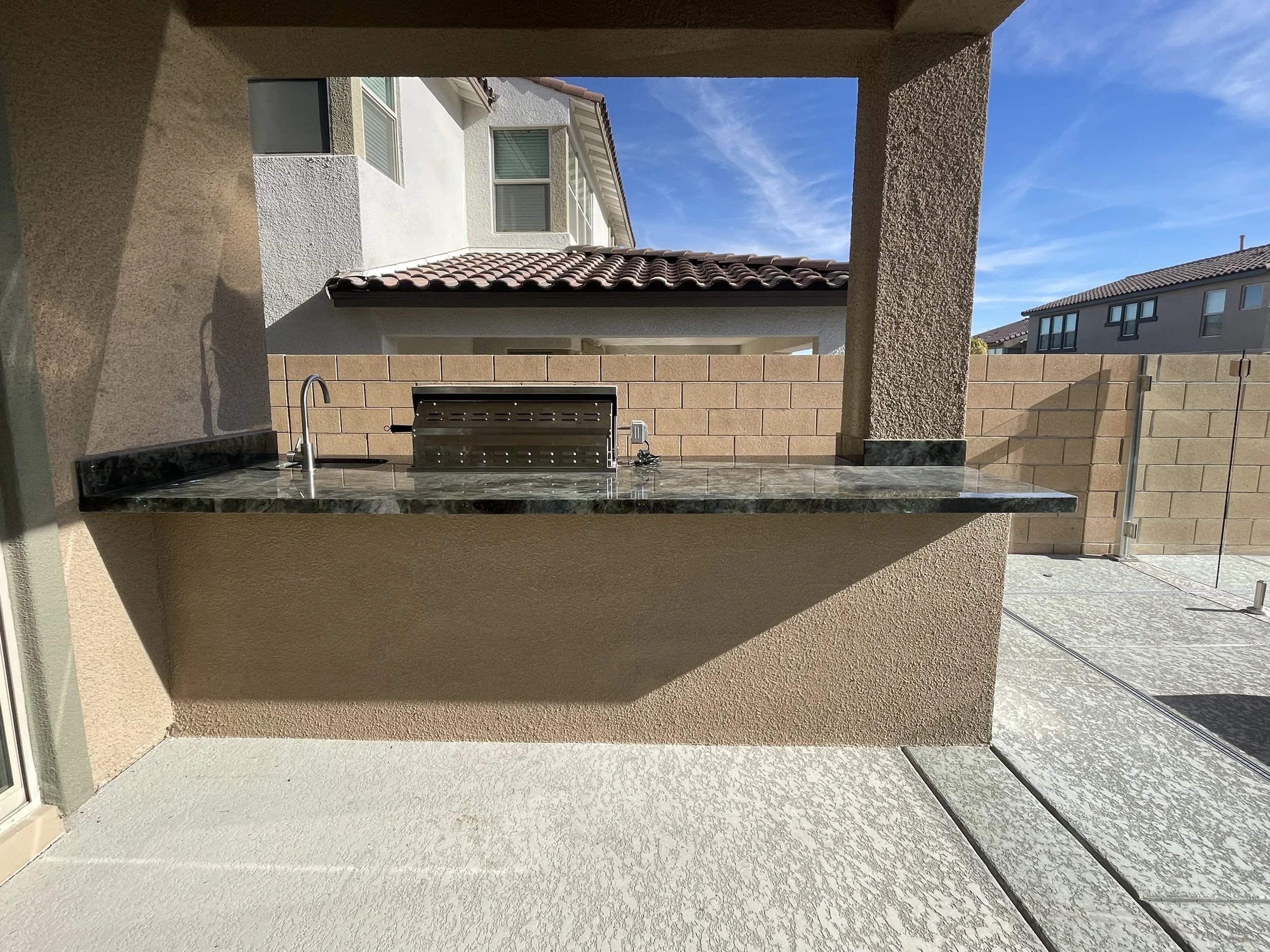 Outdoor kitchen with barbecue grill and countertop under covered patio in residential backyard with surrounding privacy wall.