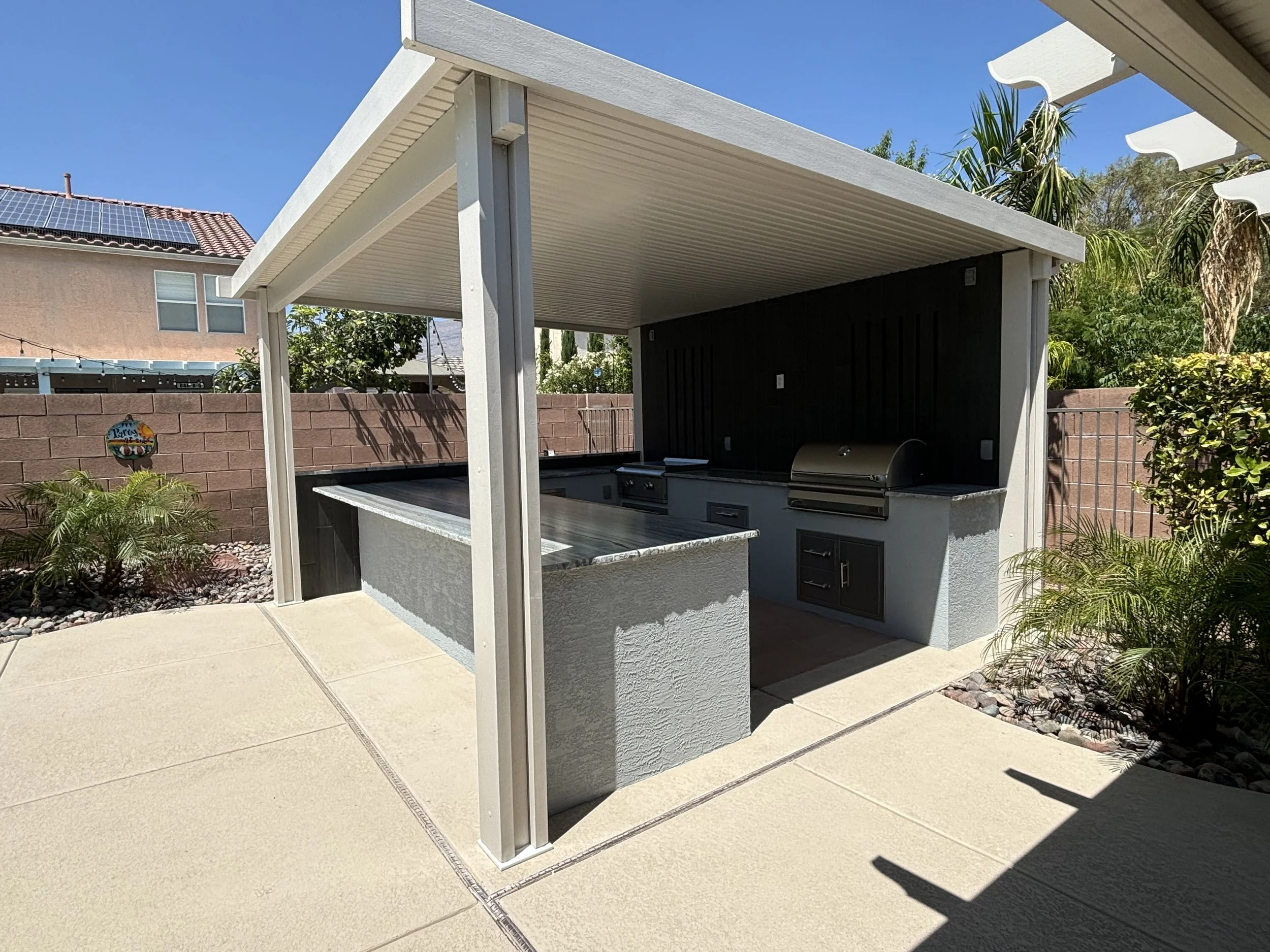 Outdoor kitchen with grill and countertop under a pergola in a backyard setting.