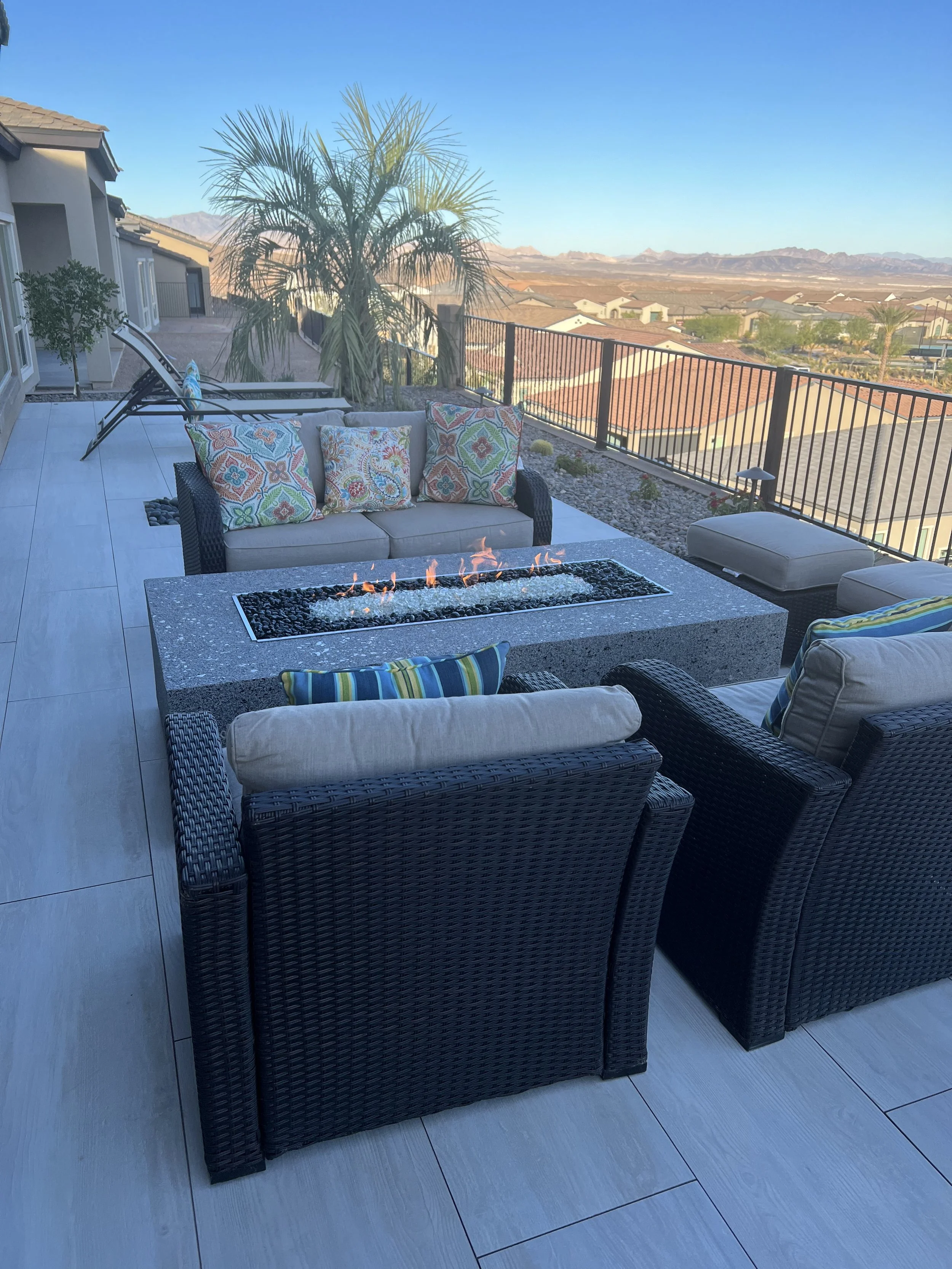 Outdoor patio with wicker chairs, decorative pillows, and a fire pit table, overlooking a scenic landscape with palm trees and mountains.