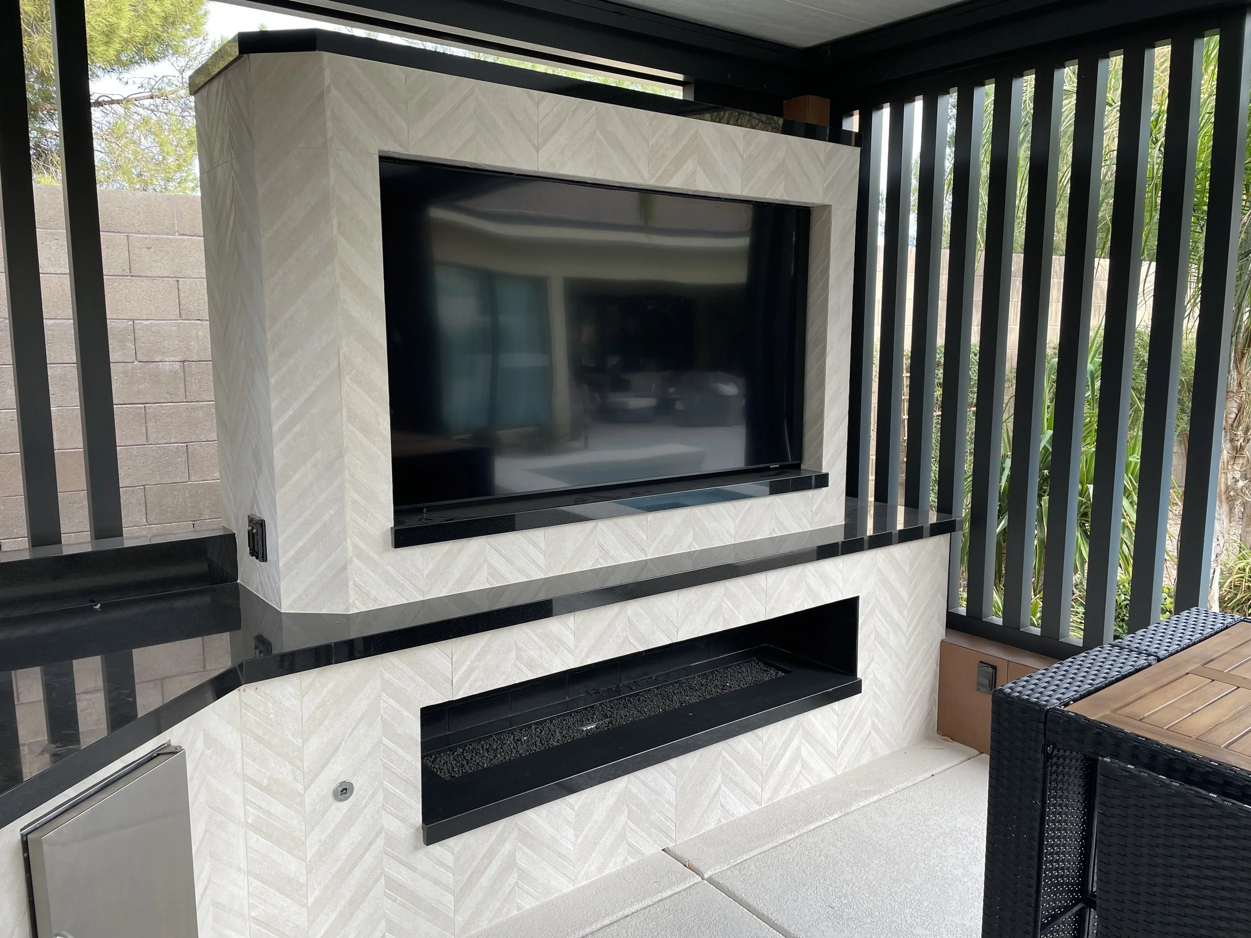 Outdoor patio with a mounted TV, modern fireplace, and wicker furniture on a tiled surface, surrounded by vertical slat walls.