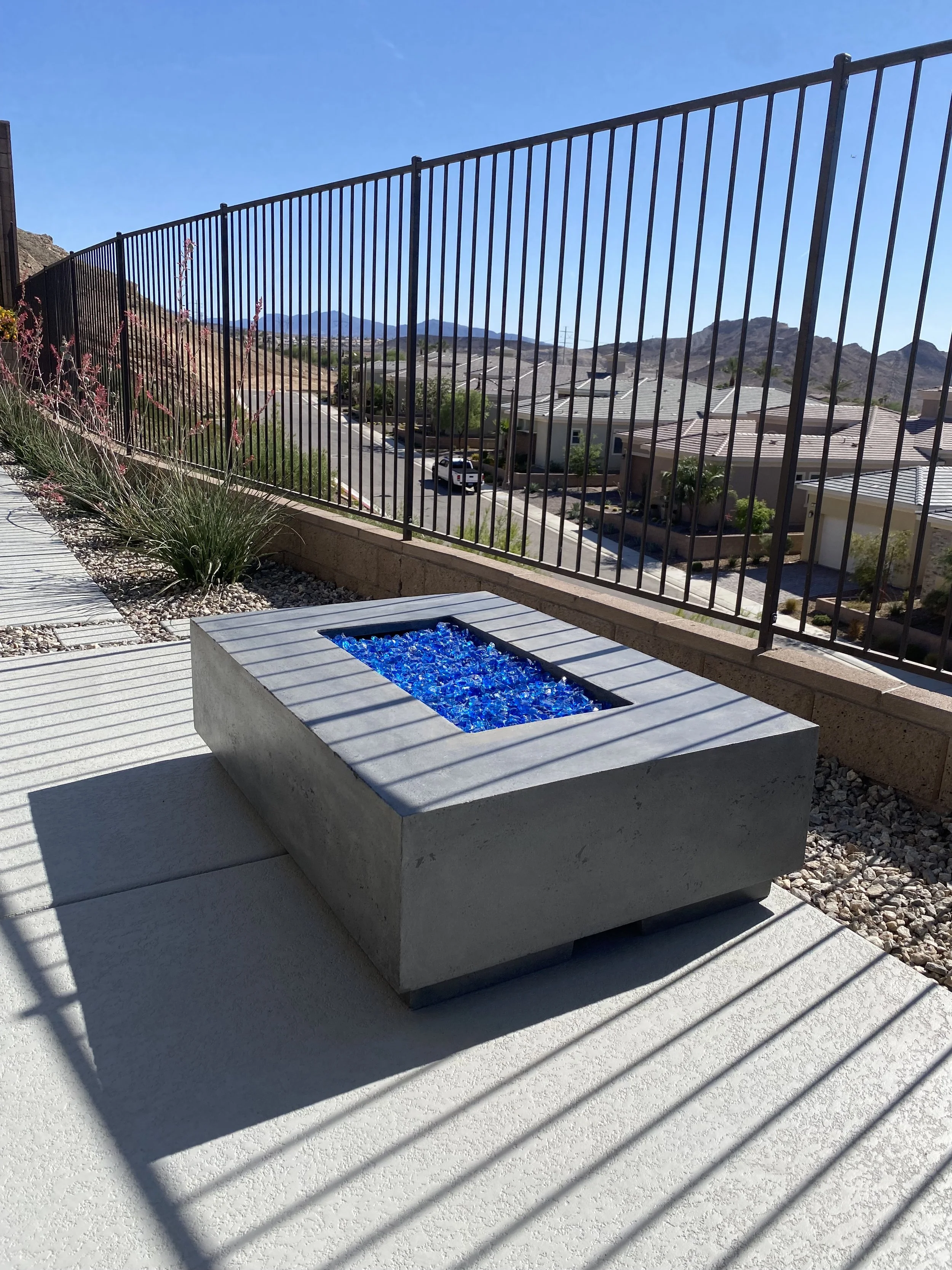 Outdoor patio with a modern square fire pit filled with blue glass stones, surrounded by a metal fence and a view of mountains and houses in the background.