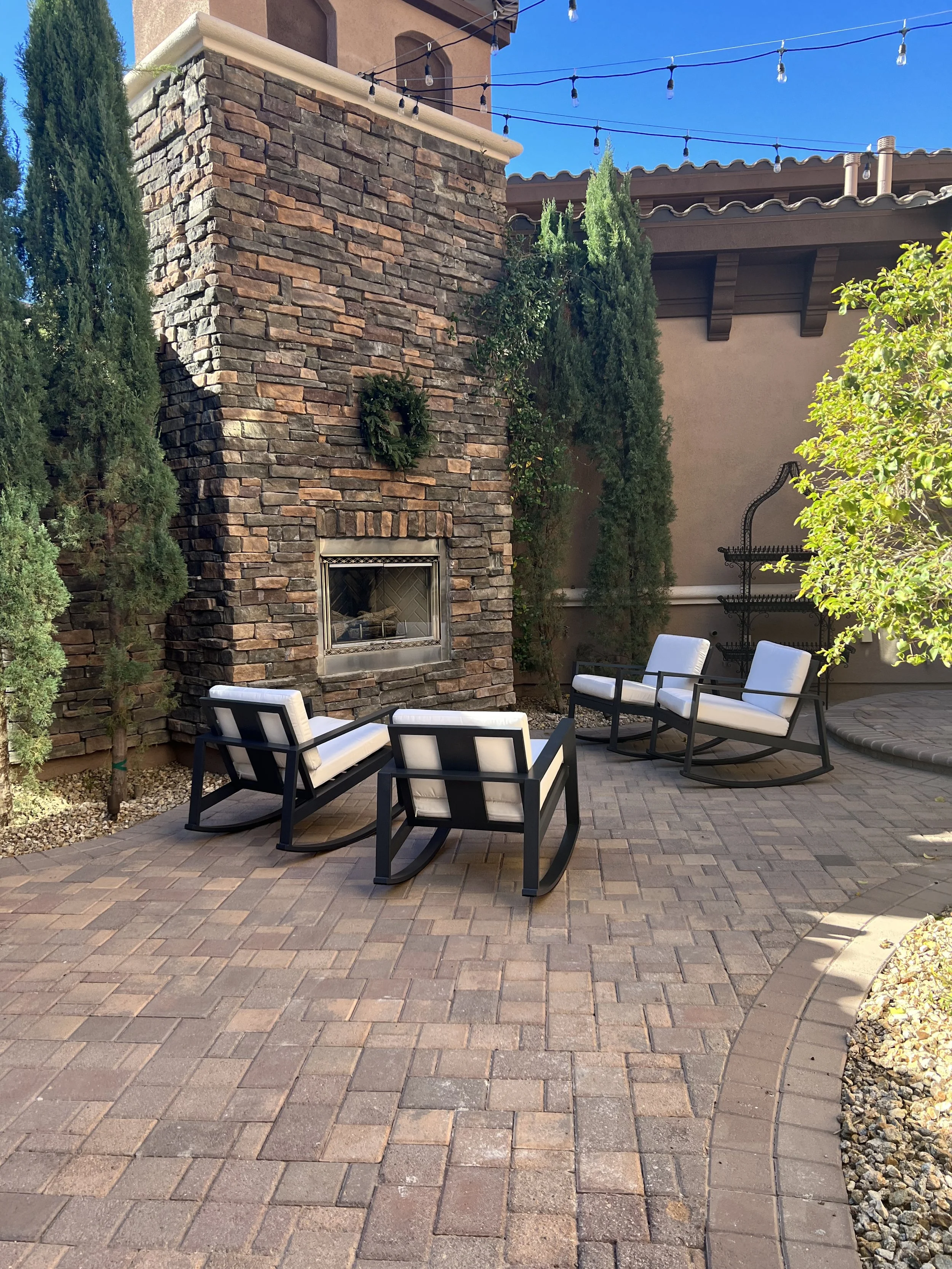 Outdoor patio with stone fireplace, surrounded by cushioned rocking chairs, string lights above, and potted trees and plants.