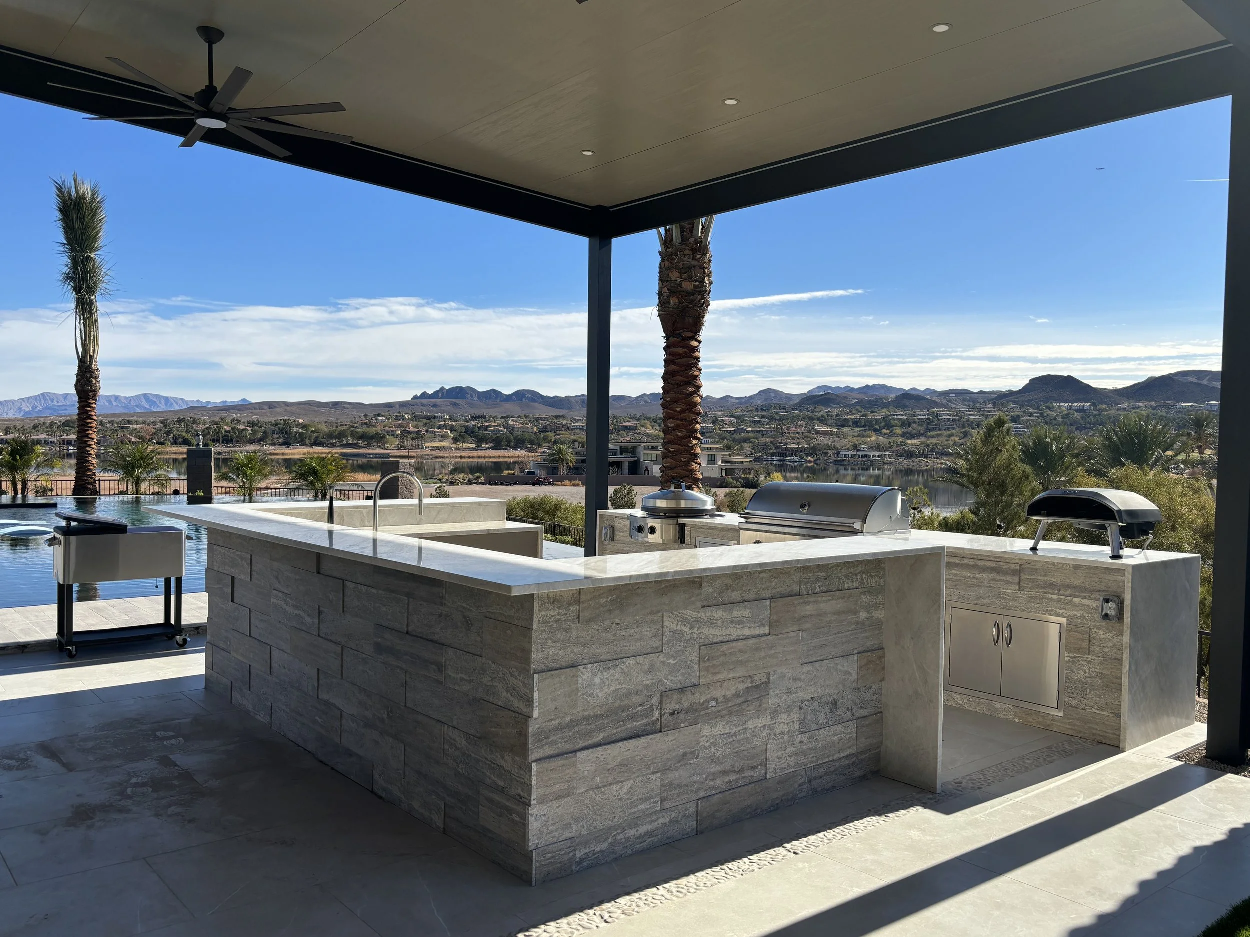Outdoor kitchen with a modern design featuring stone countertops, built-in grill, and pizza oven under a shaded patio with a scenic mountain and palm tree view.