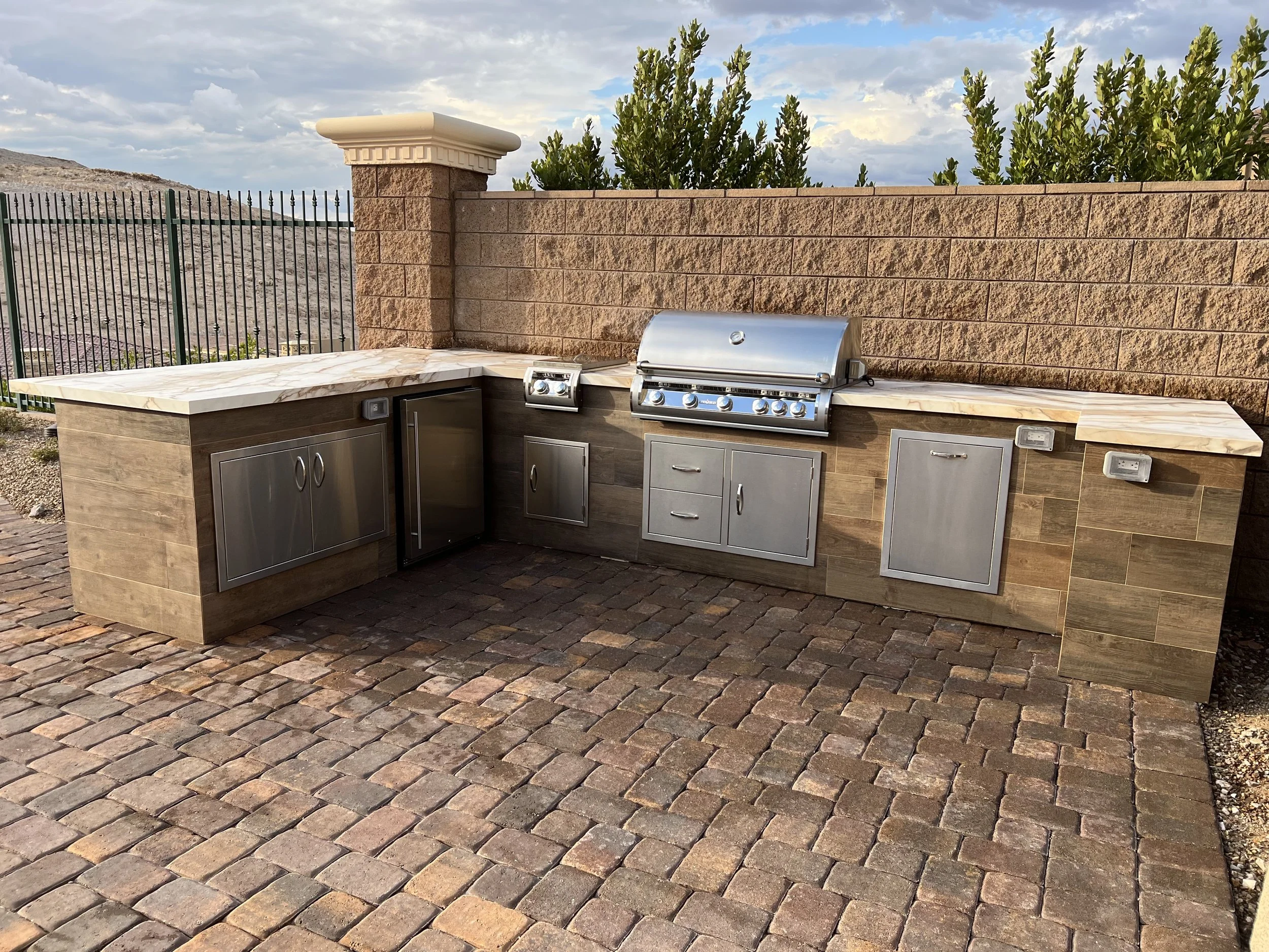 Outdoor kitchen with stainless steel grill and storage cabinets, on a brick patio.