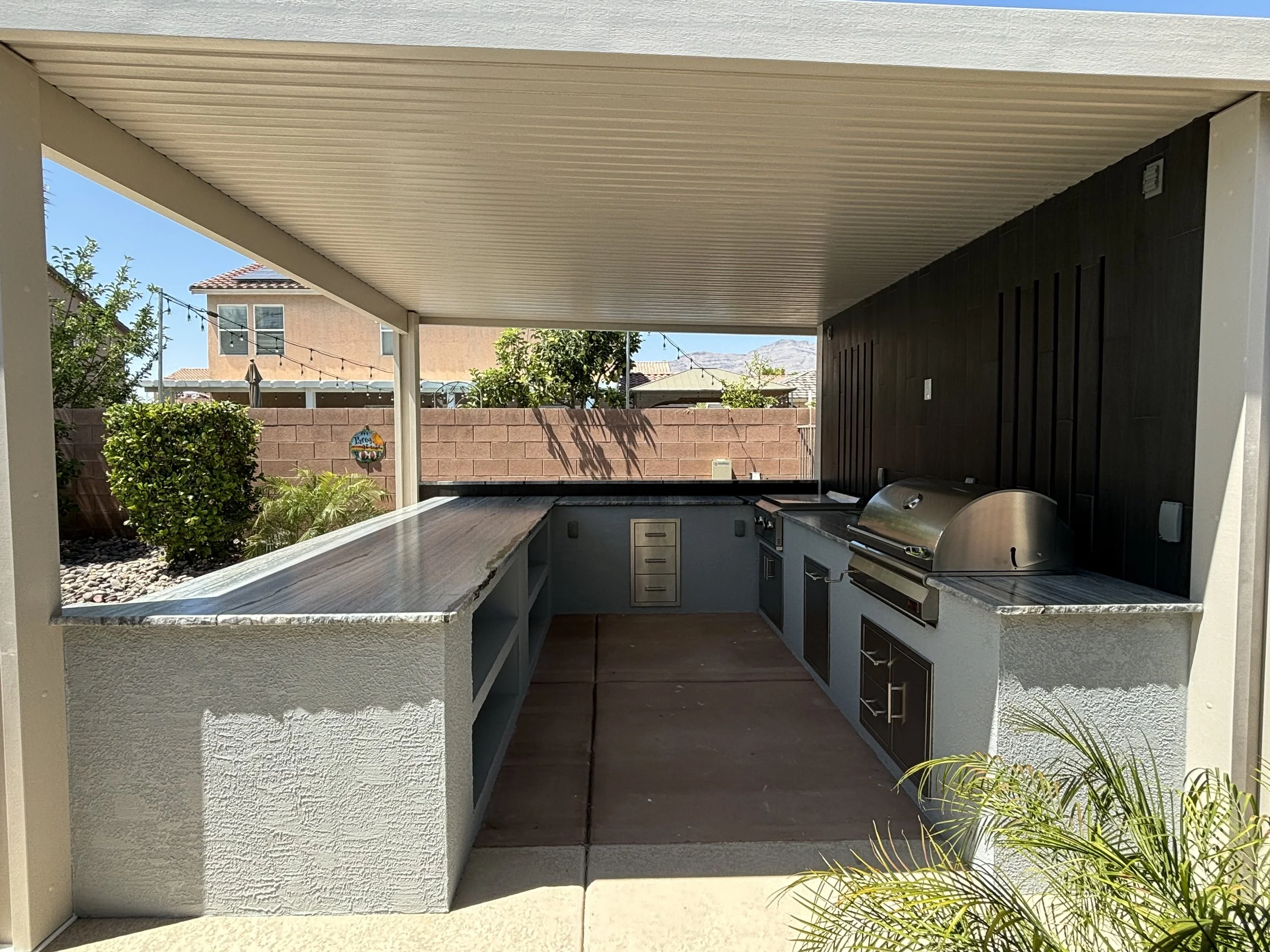 Outdoor kitchen with a covered patio, featuring a built-in grill, countertop, storage drawers, and a landscaped backyard.
