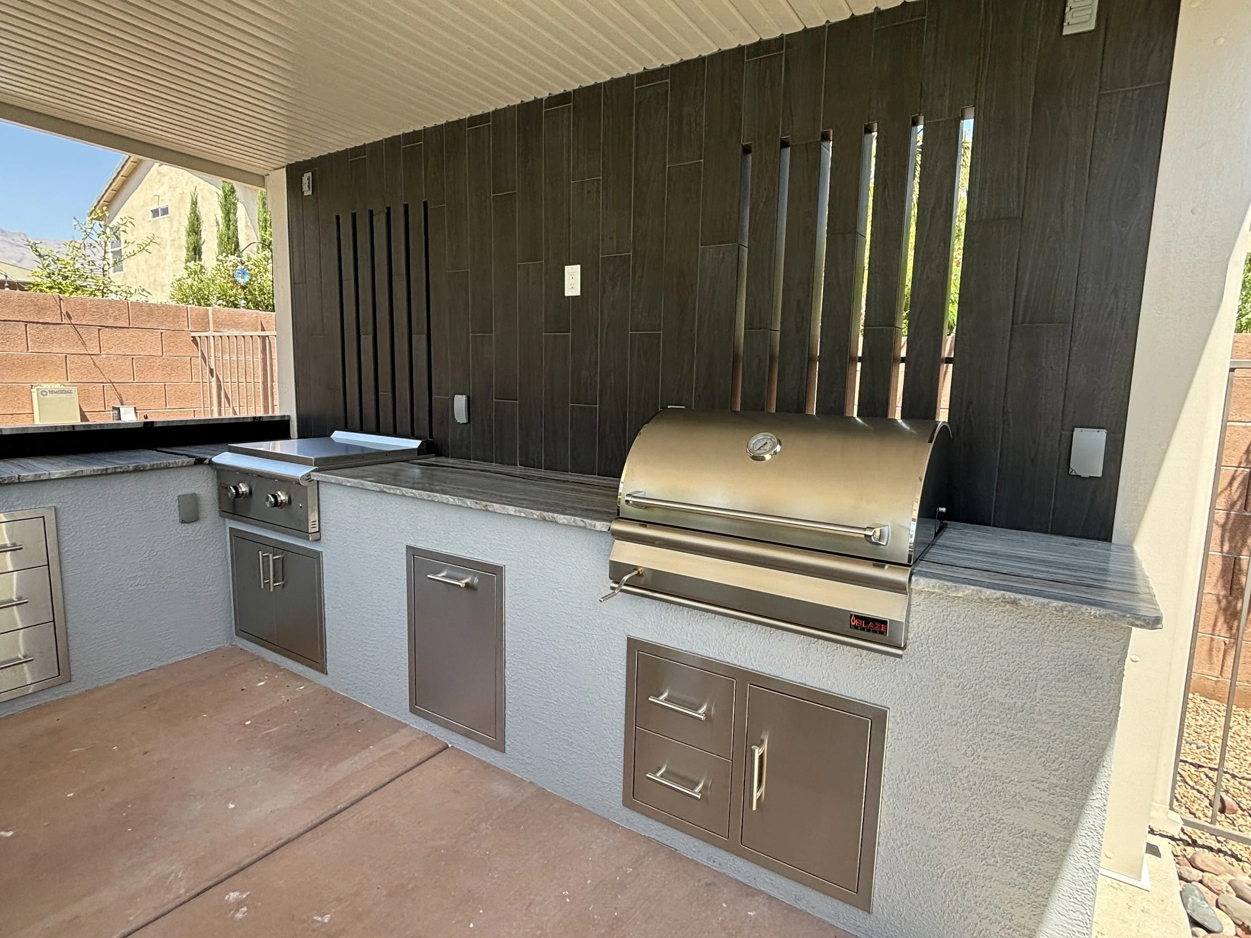 Outdoor kitchen with grill, side burner, sink, and storage cabinets set against a tiled backsplash.