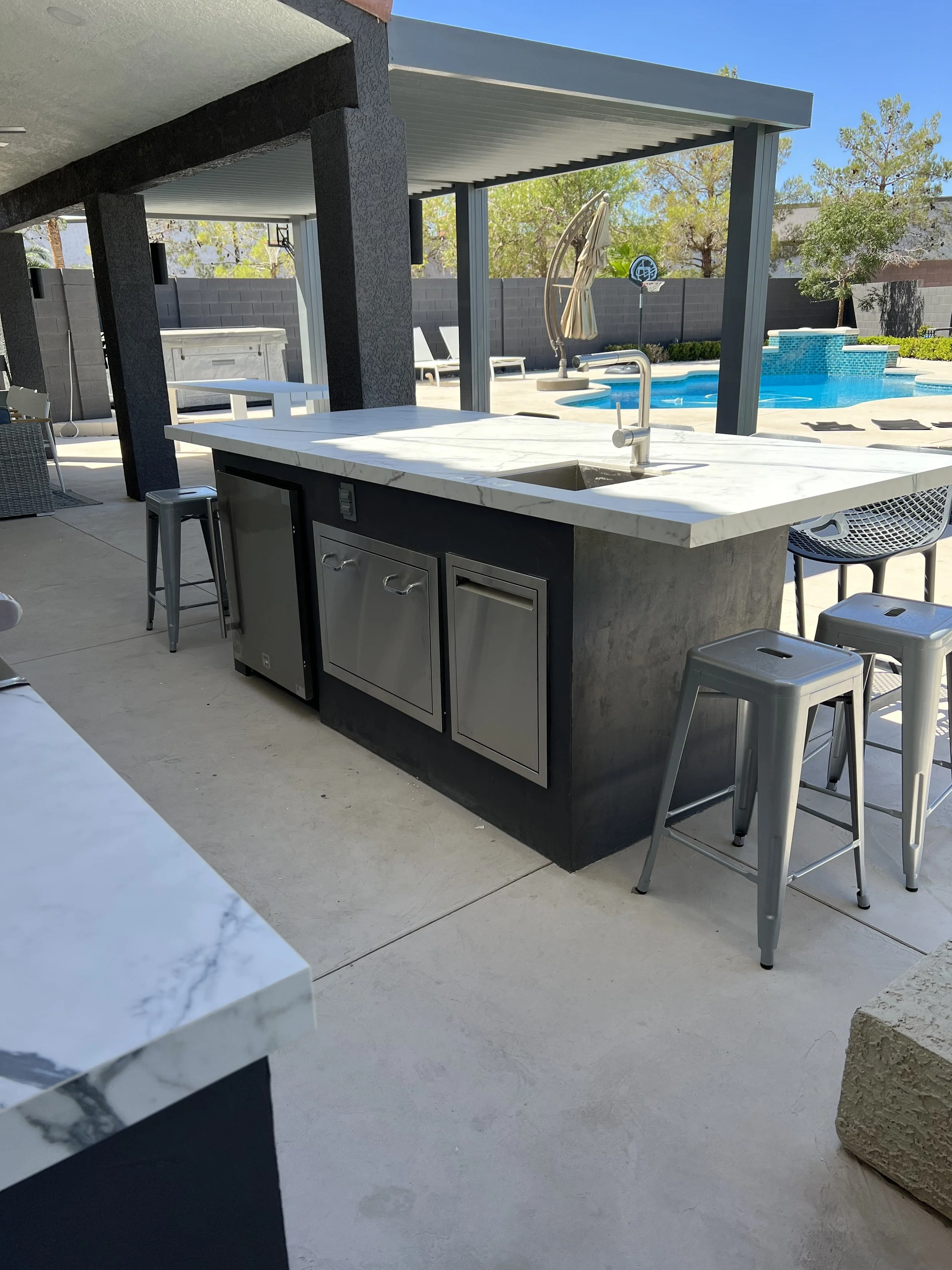 Outdoor kitchen with a marble countertop, stainless steel appliances, a sink, metal bar stools, and a covered patio. Background features a swimming pool, lounge chairs, and a fenced yard.