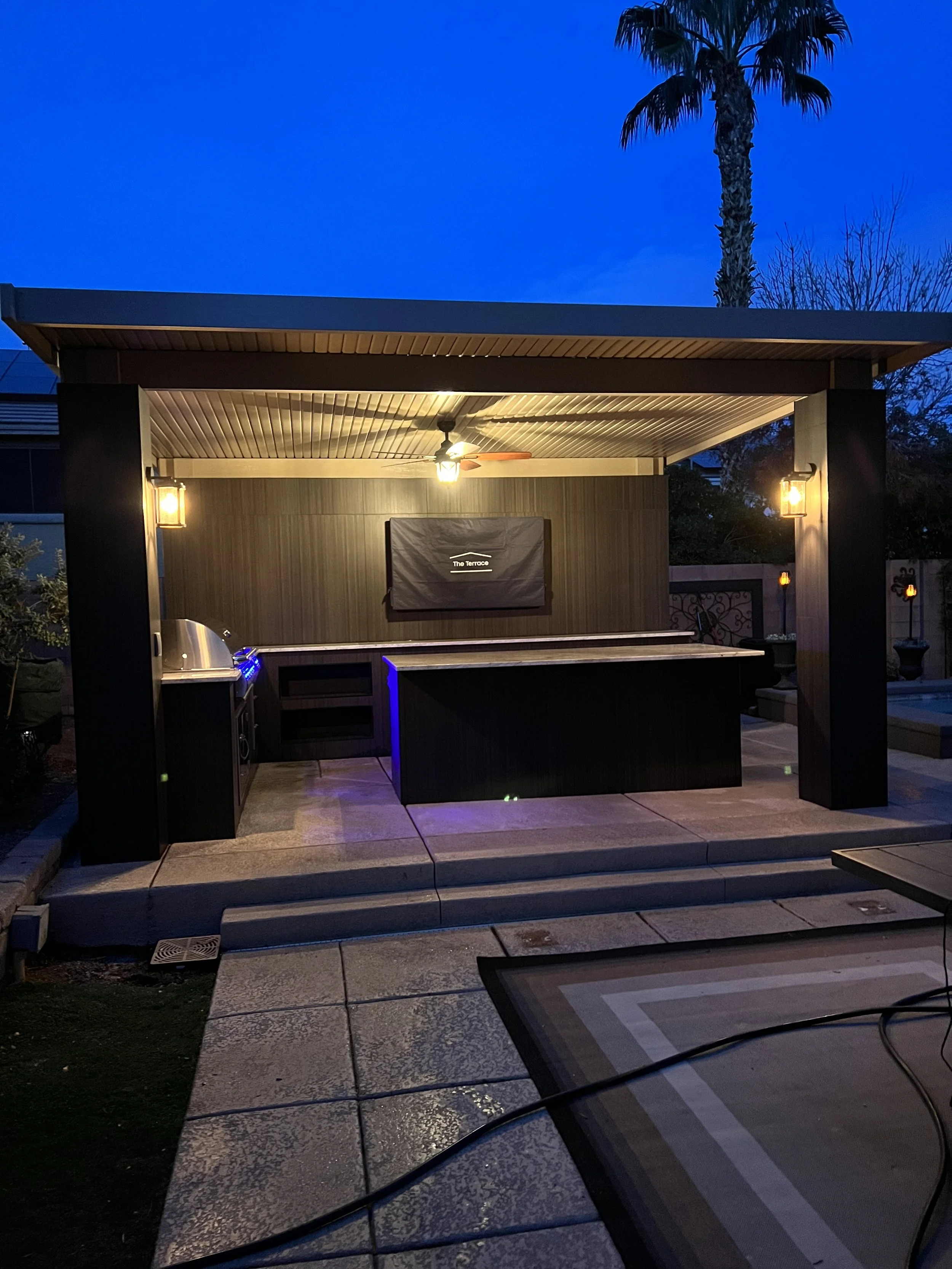 Outdoor kitchen with grill under a gazebo at dusk, featuring lighting and a ceiling fan.