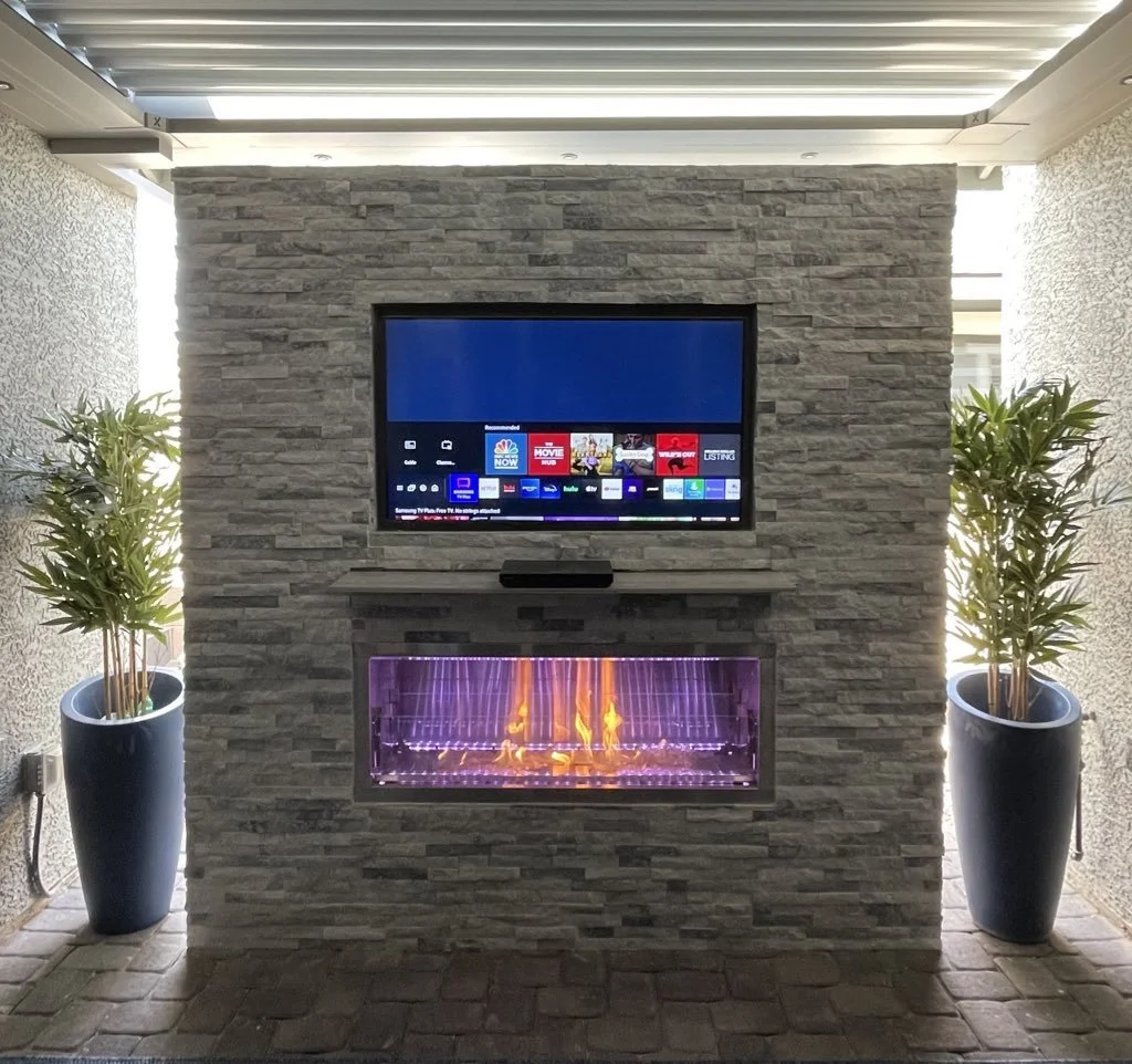 Outdoor stone wall with a mounted flat-screen TV and electric fireplace, surrounded by two potted plants.