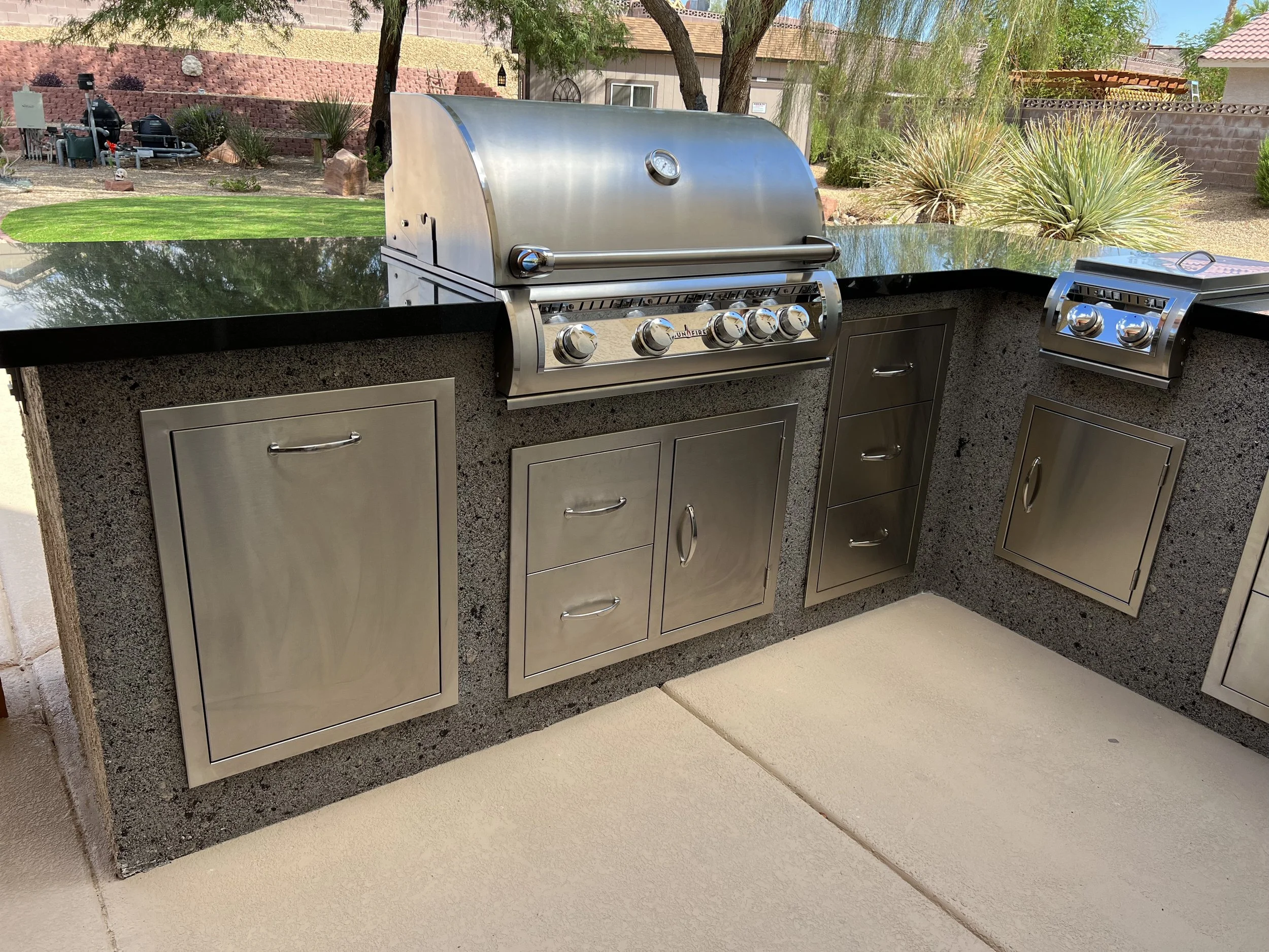 Outdoor kitchen island with stainless steel grill, storage drawers, and cabinets on a concrete patio.