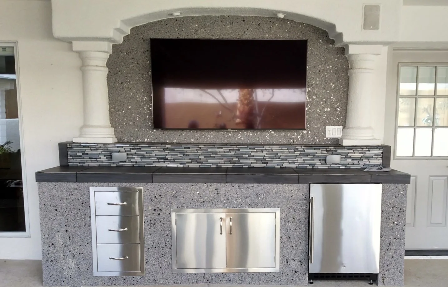 Outdoor kitchen with granite countertops, built-in stainless steel appliances, a large mounted TV, and decorative tile backsplash.
