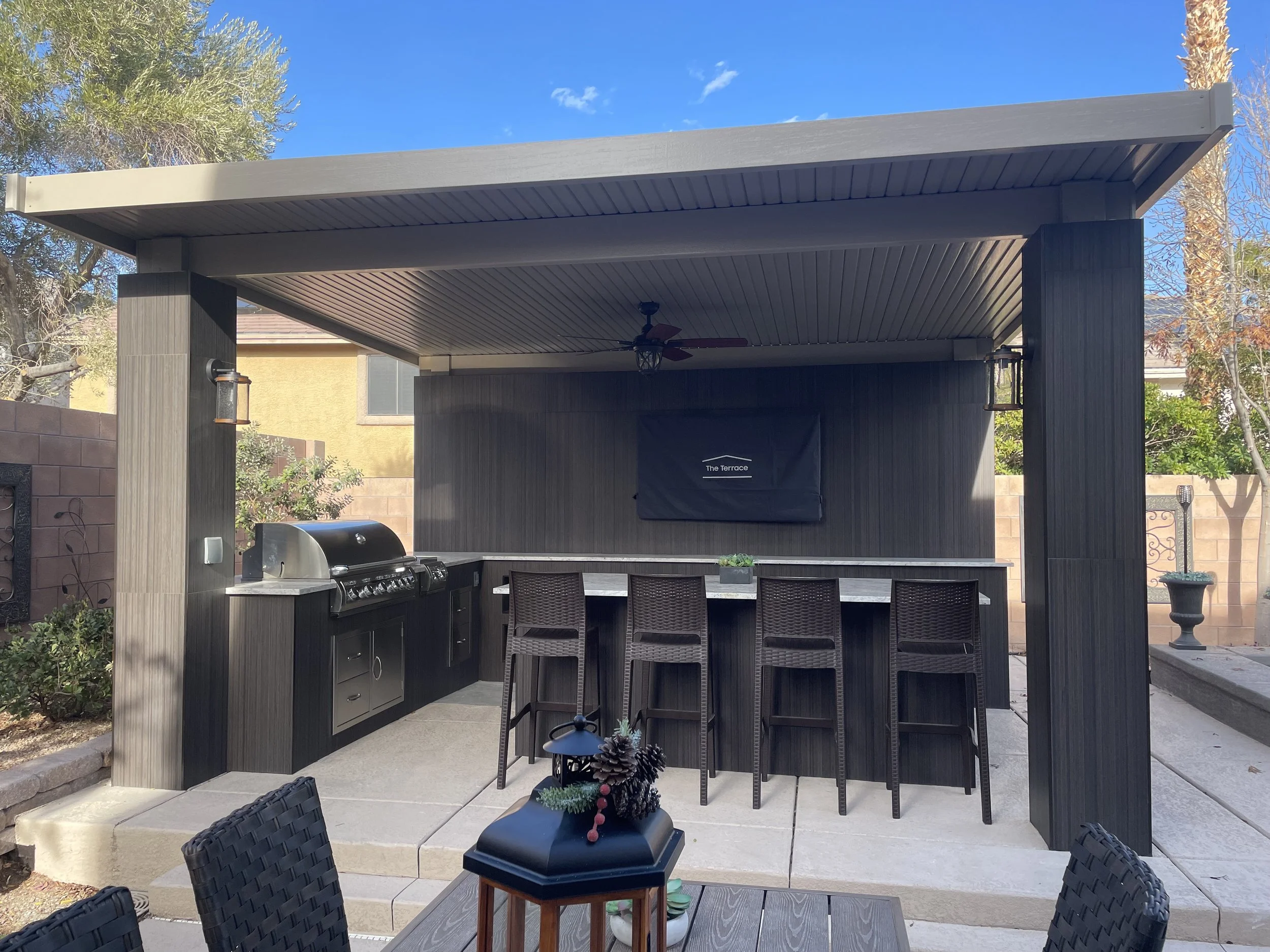 Outdoor kitchen area with a covered pergola featuring a large grill, countertop, and bar seating with several stools. The space is surrounded by a patio with additional seating and greenery.