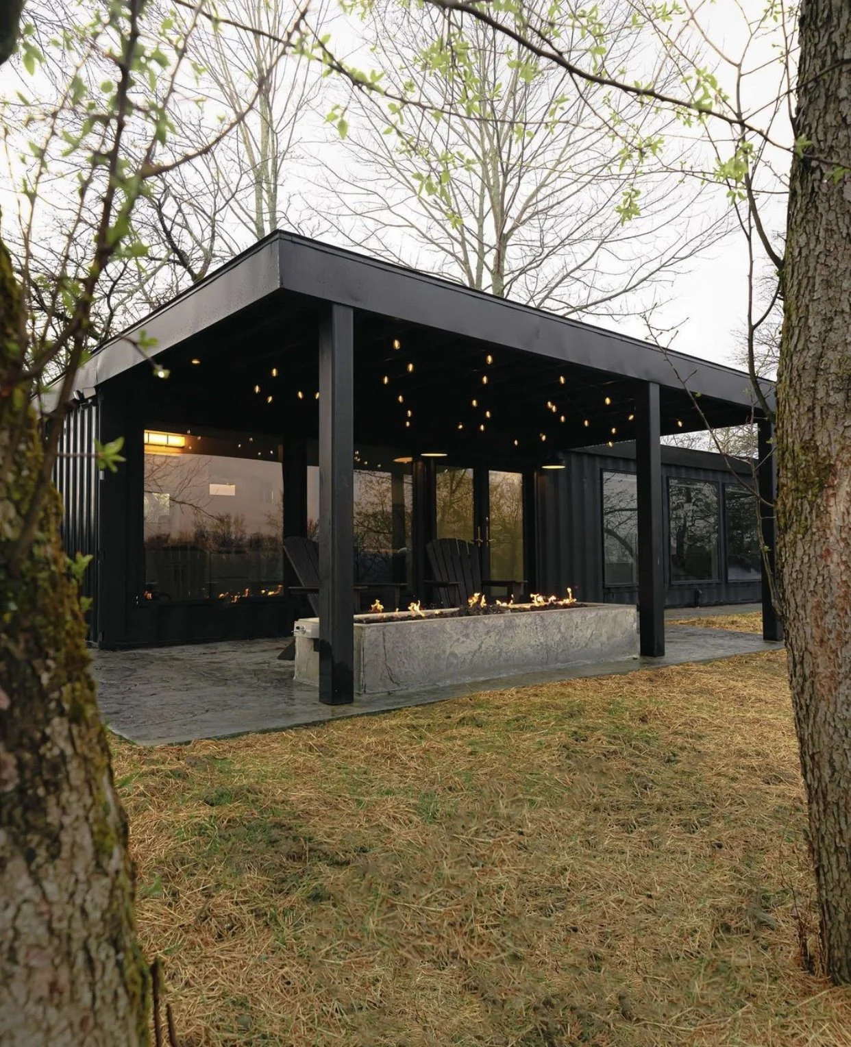 Modern black outdoor pavilion with string lights, surrounded by trees and dry grass.