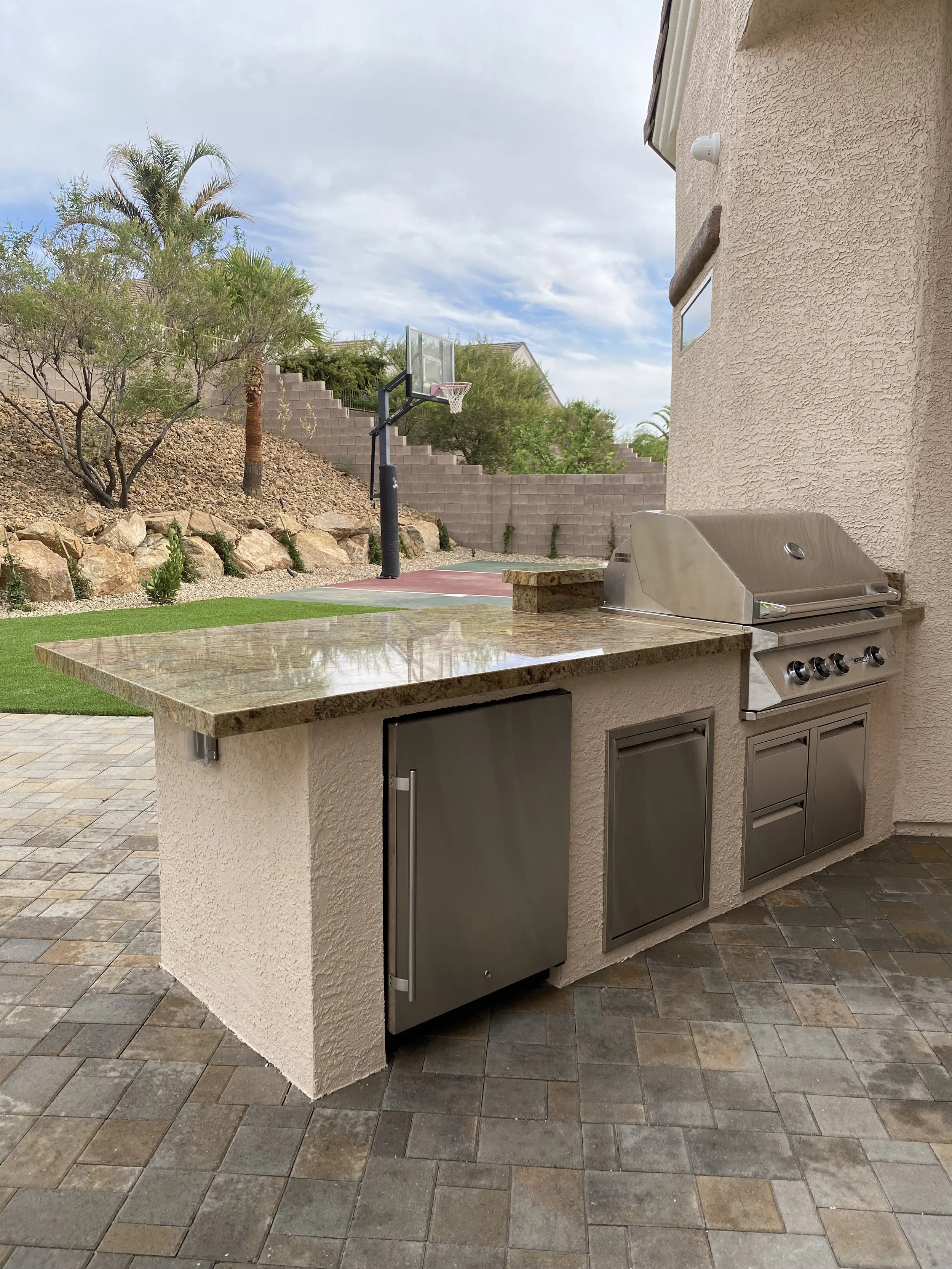 Outdoor patio with built-in kitchen, featuring a stainless steel grill, fridge, and storage. Nearby is a basketball hoop, surrounded by landscaped garden with trees and rocks.