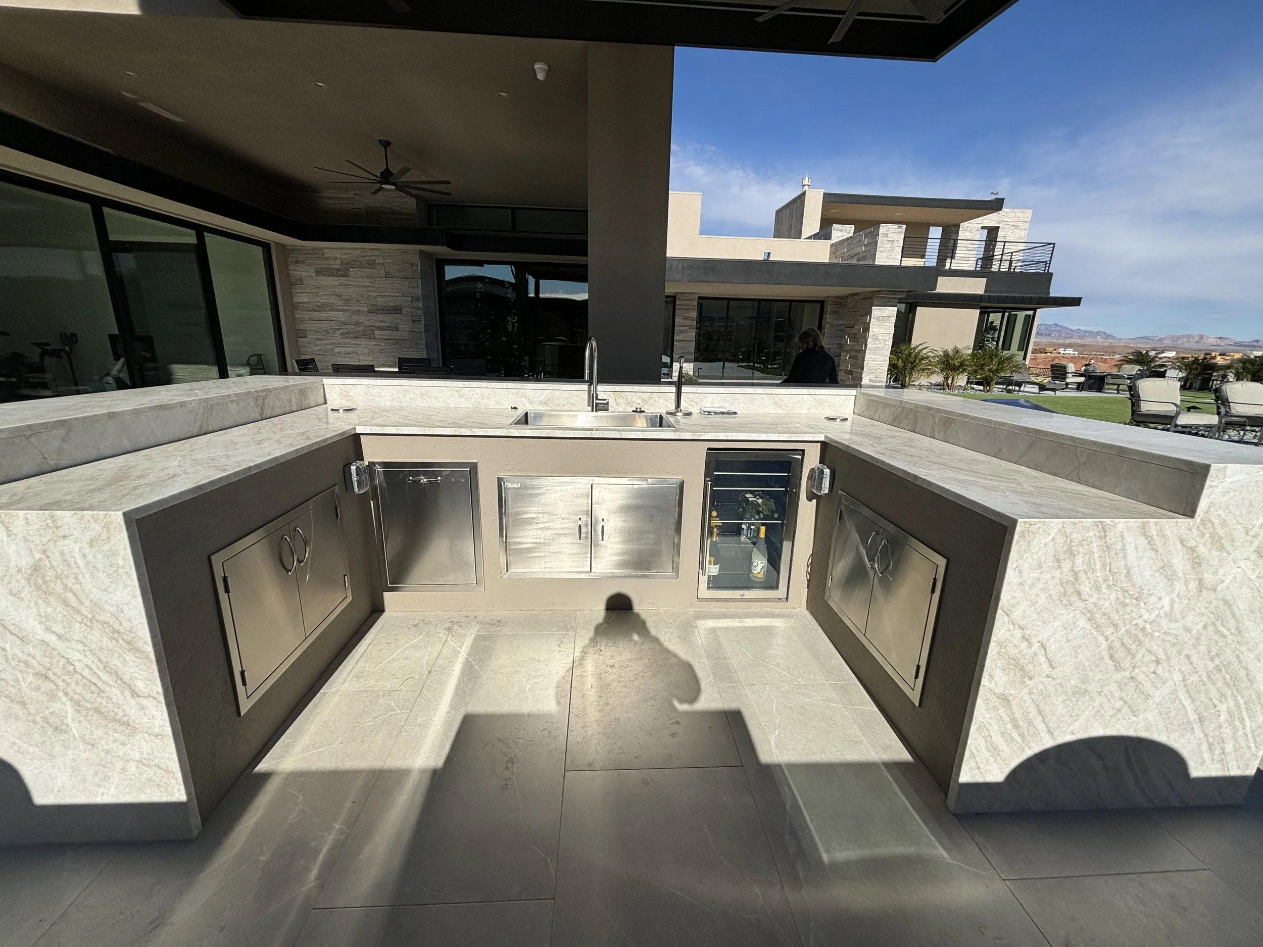 Outdoor modern kitchen with stone countertops, stainless steel appliances, and a sink, set against a contemporary house with large windows and a patio.