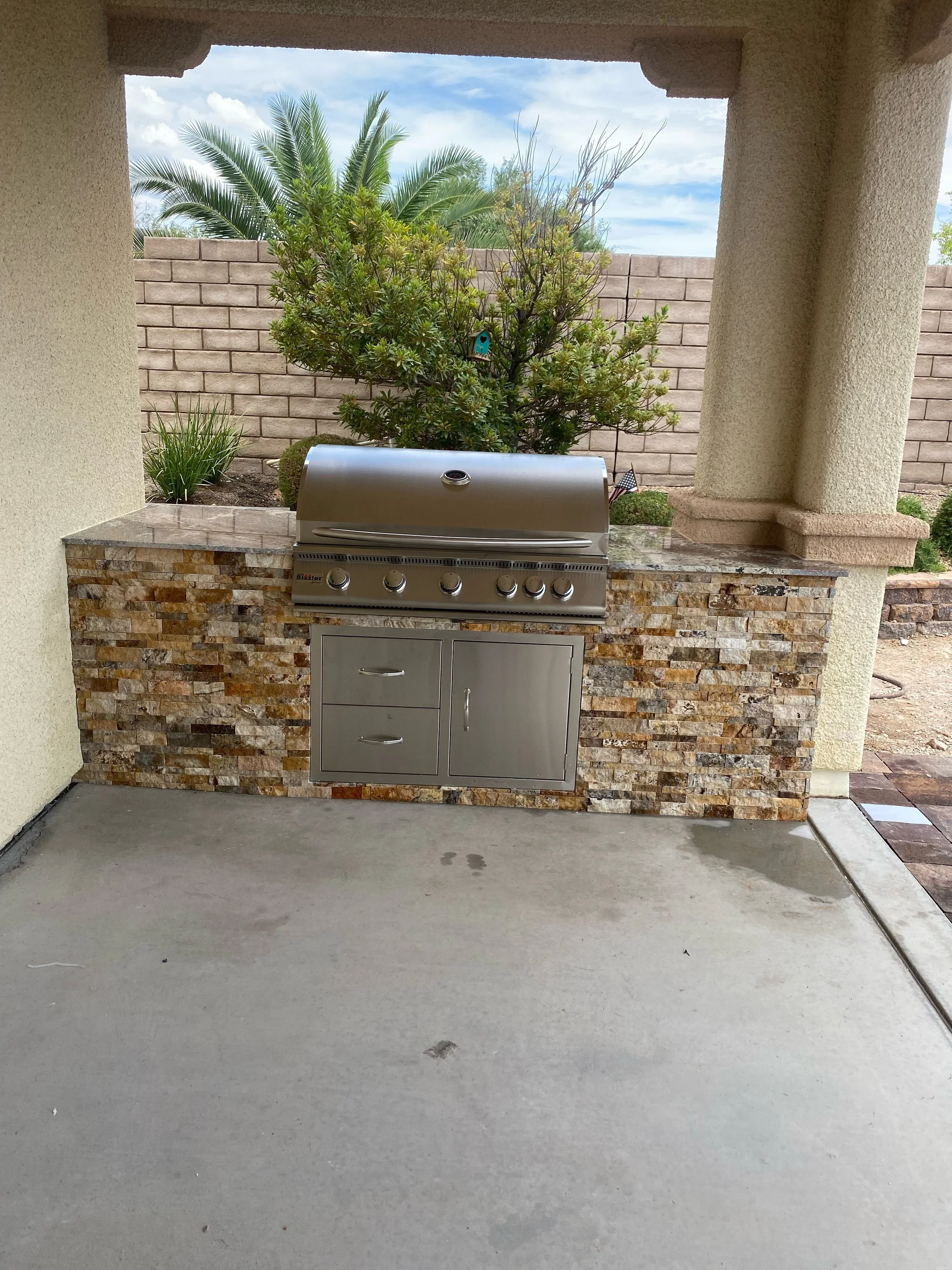 Outdoor built-in grill with stone surround under a covered patio.