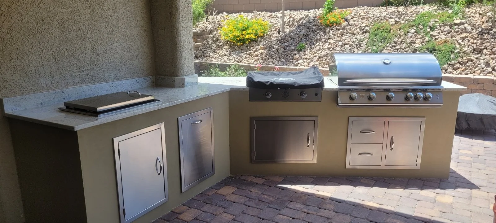 Outdoor kitchen with built-in grill, countertop, and storage cabinets on a patio, surrounded by a garden with rocks and plants.