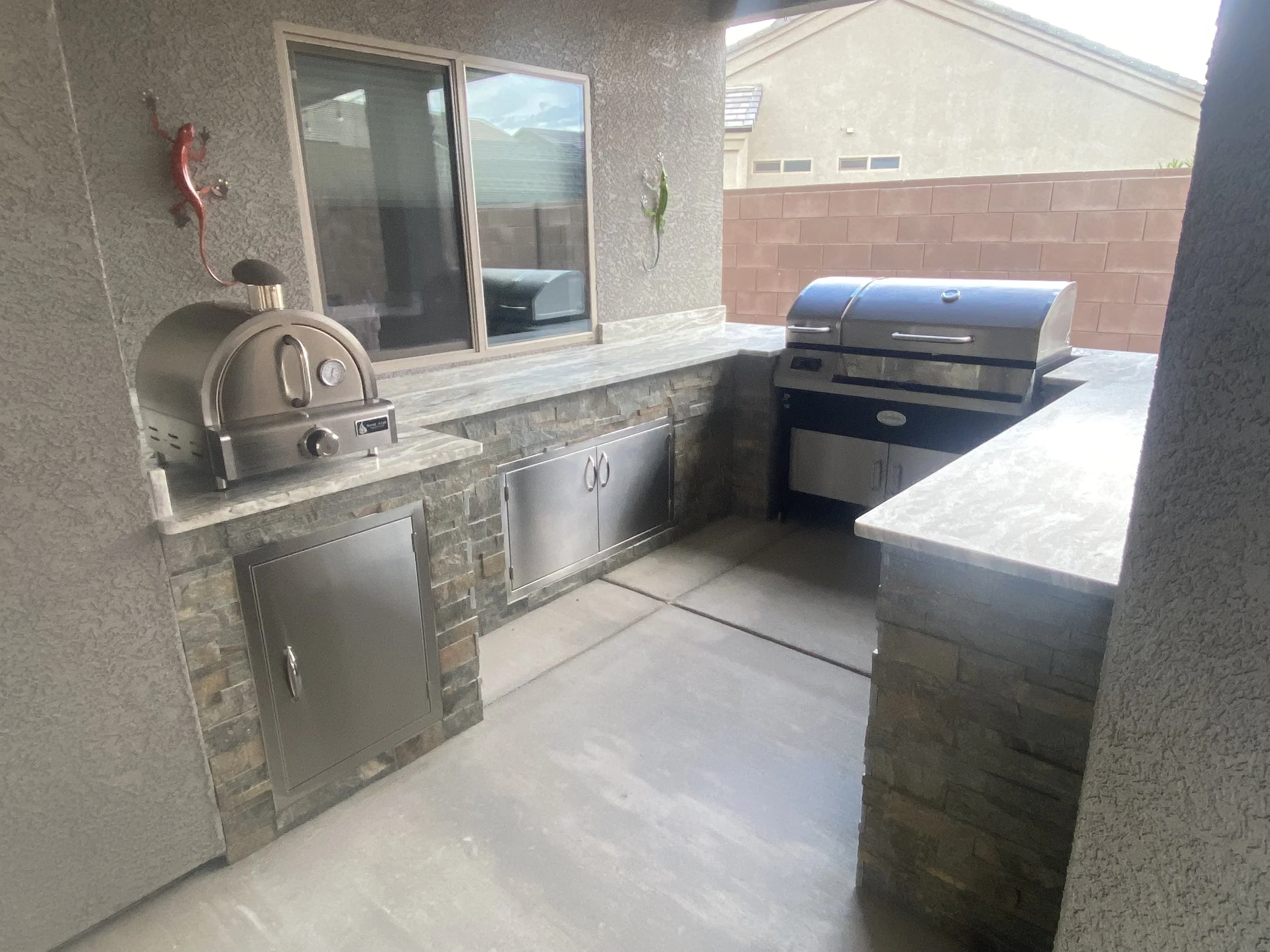 Outdoor kitchen with stone countertops, a grill, pizza oven, and storage cabinets against a stucco wall with decorative geckos.