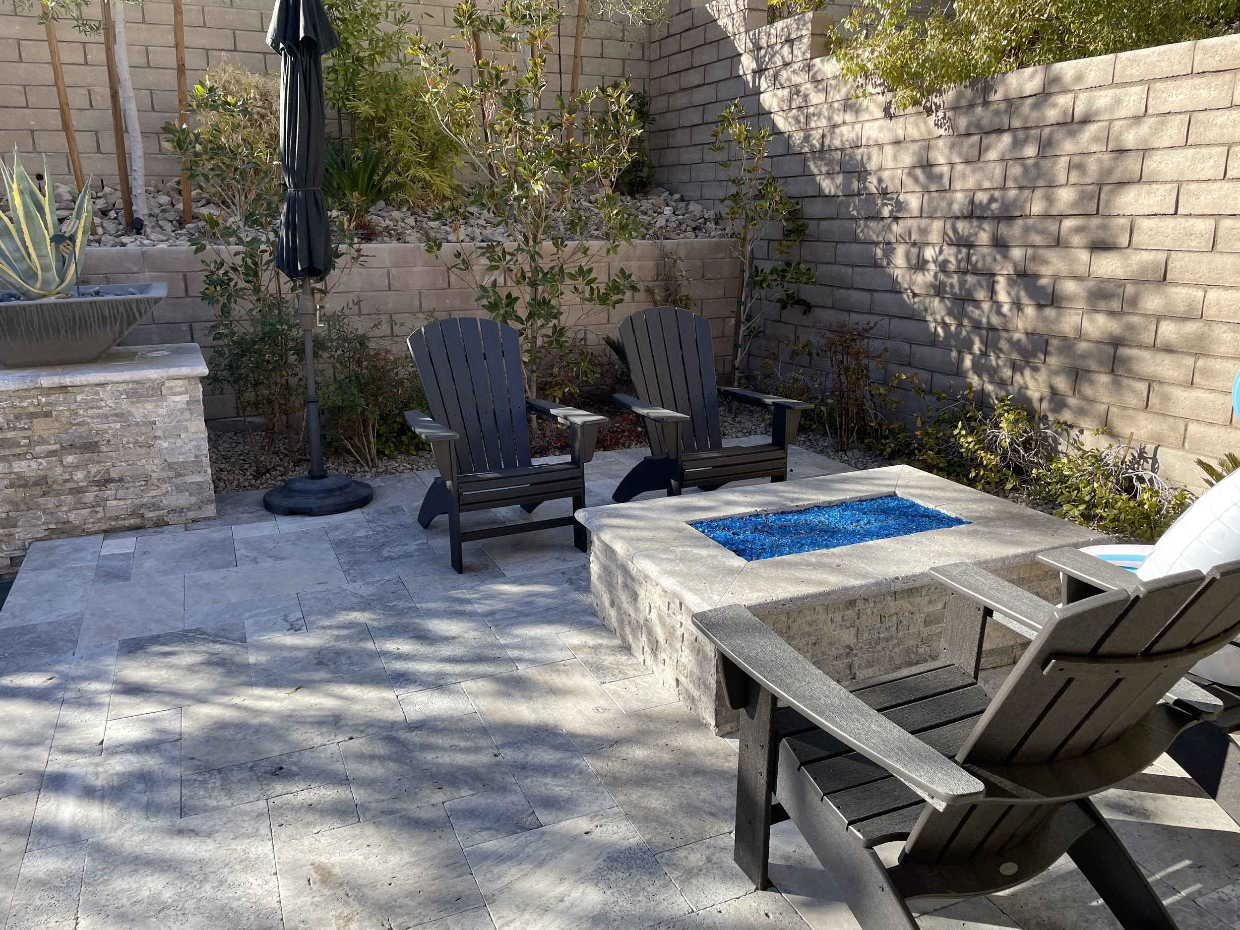 Outdoor tiled patio with black Adirondack chairs surrounding a stone fire pit filled with blue glass chips; stone wall and greenery in the background.