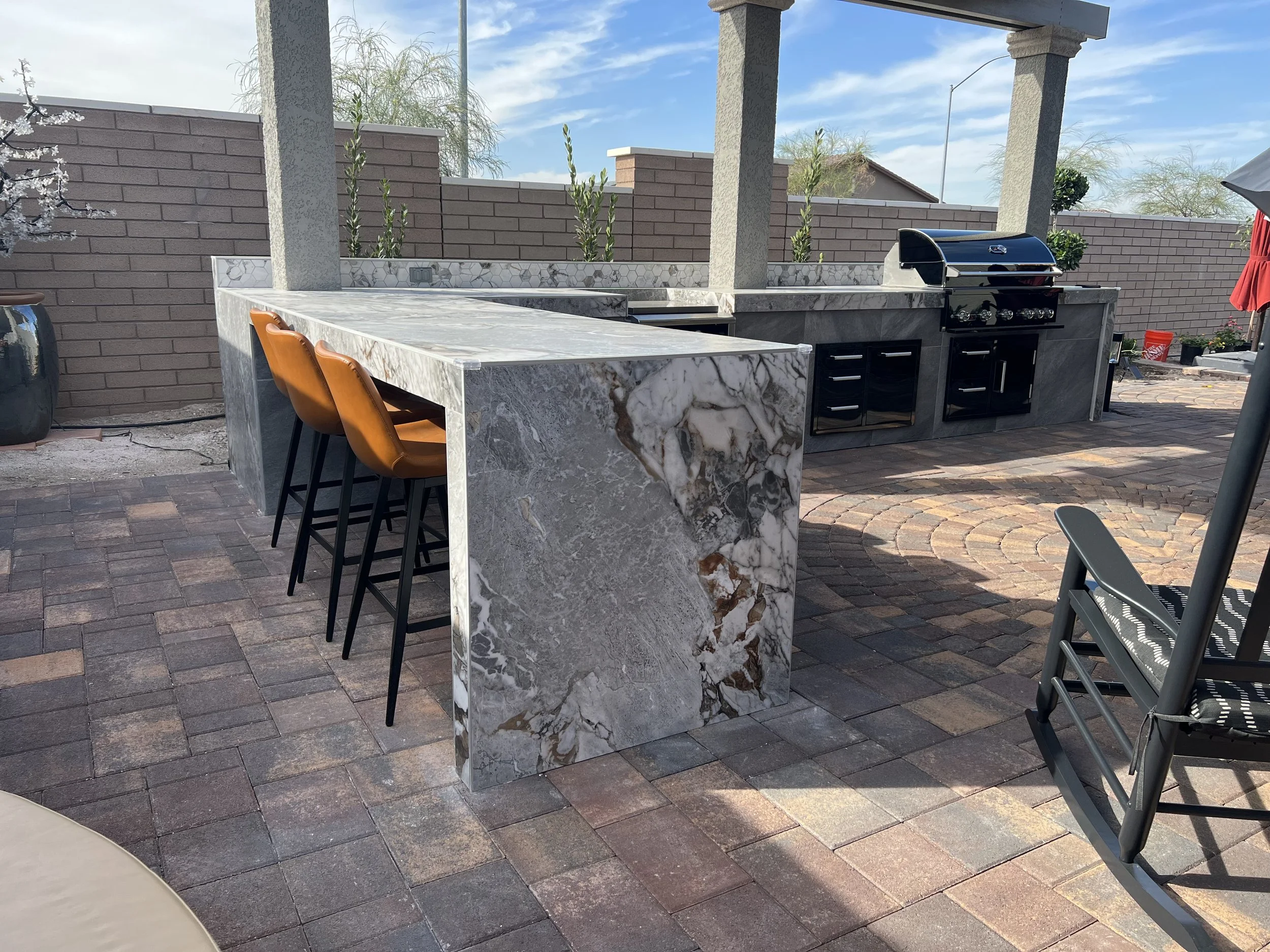 Outdoor kitchen area with marble countertops, bar stools, and a built-in grill on a brick patio.