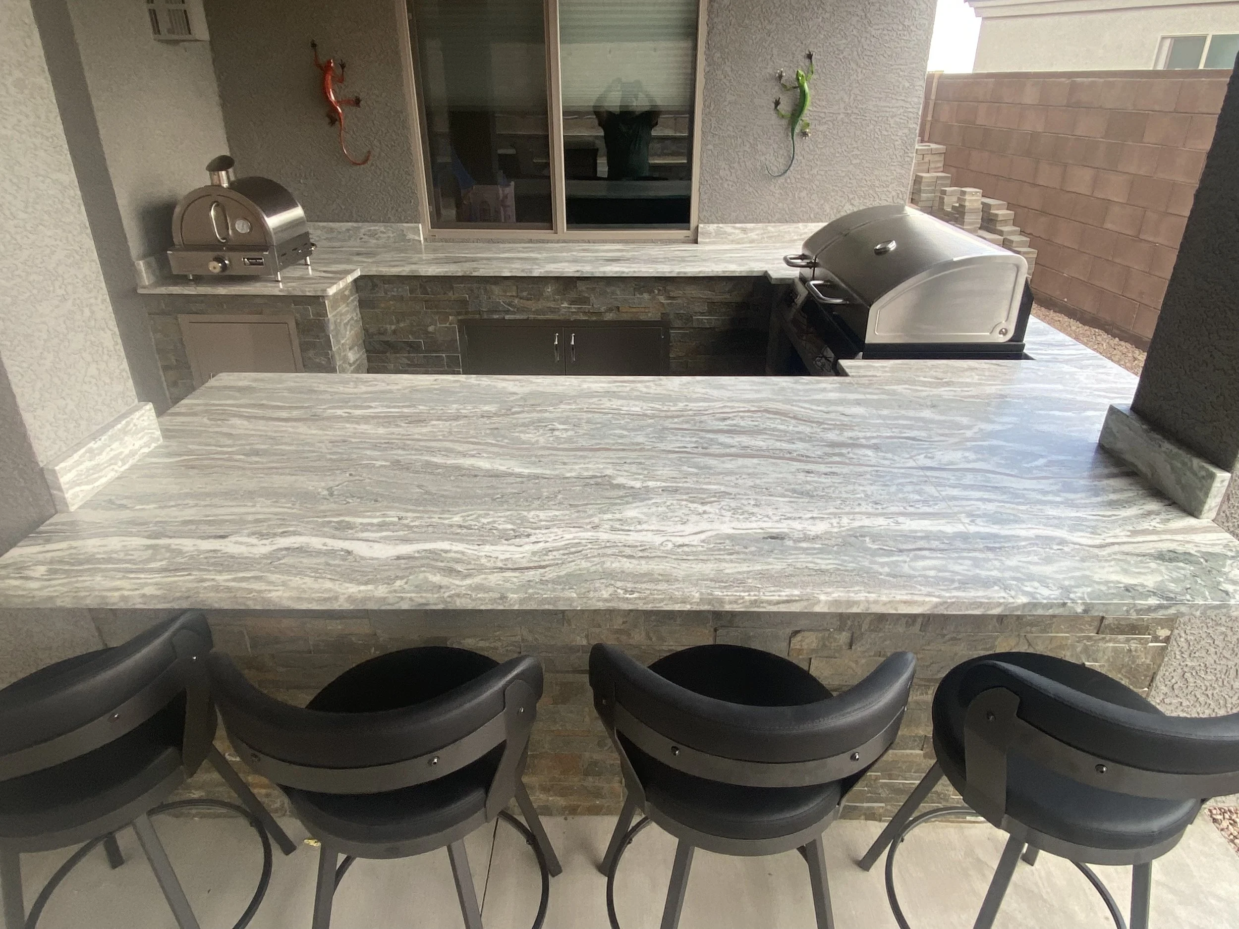 Outdoor kitchen with a marble countertop, bar stools, pizza oven, and gas grill.