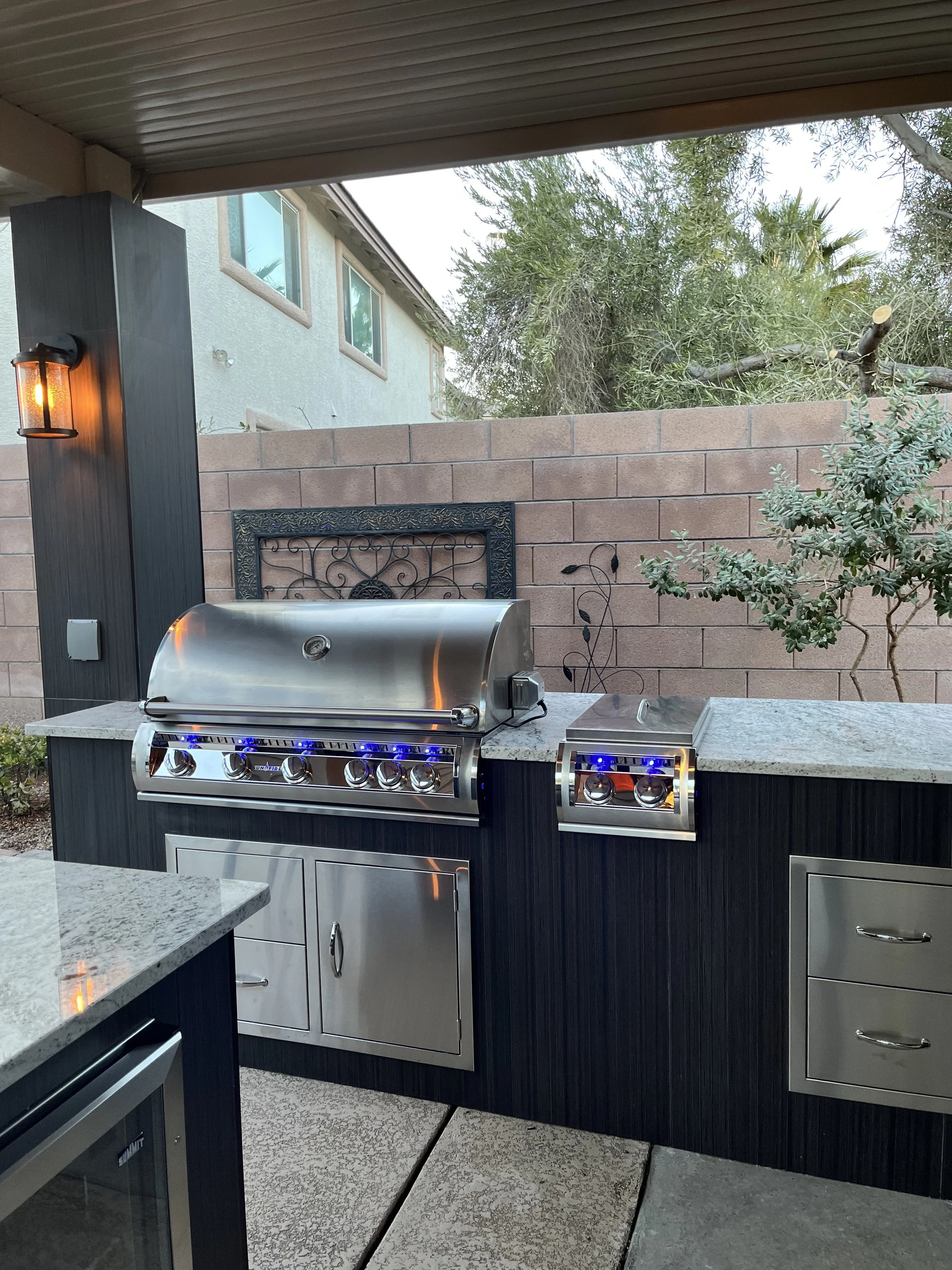Outdoor kitchen with a stainless steel gas grill, granite countertops, and cabinetry, under a covered patio. A stone wall and tree are visible in the background.