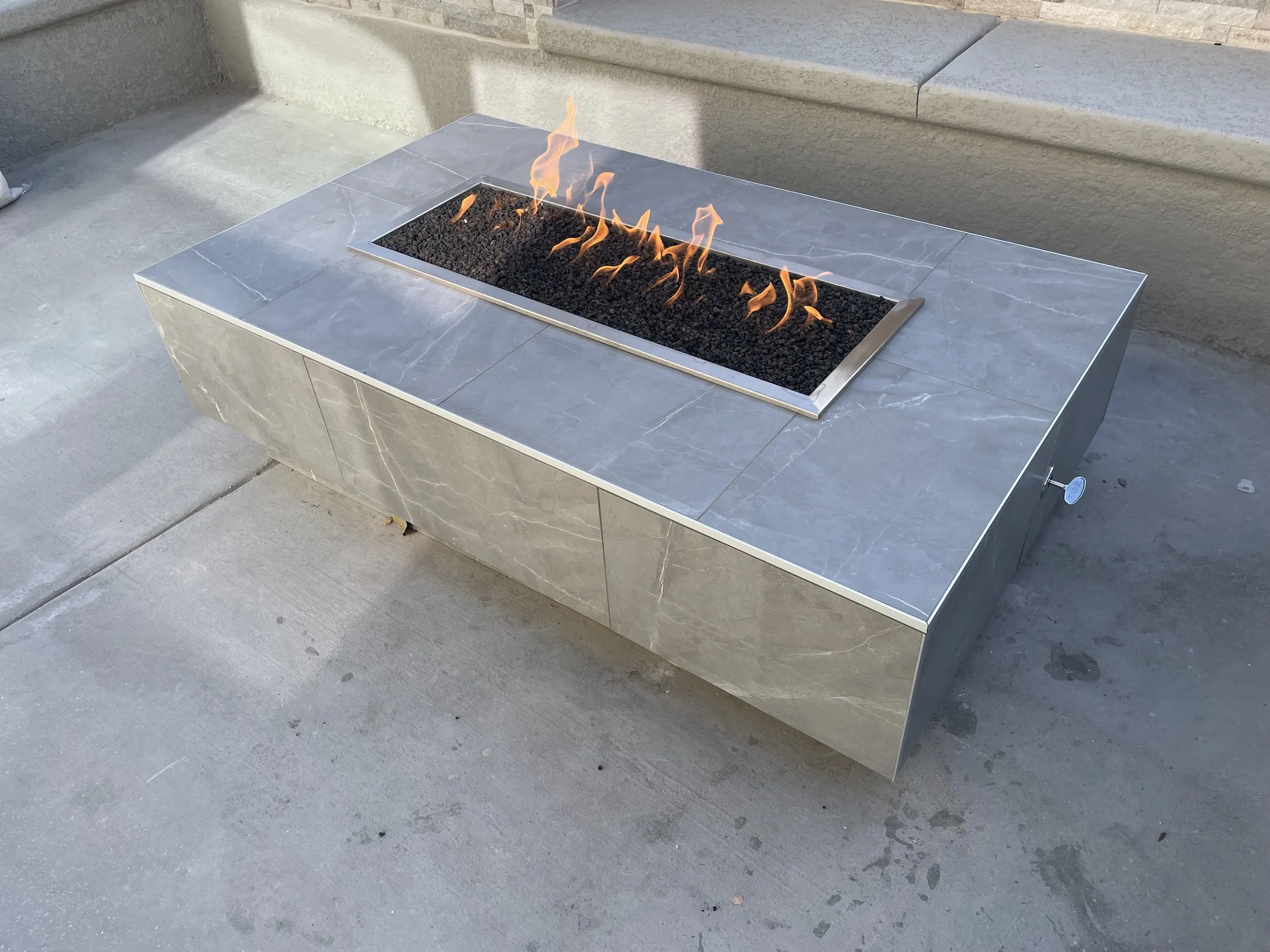 Modern outdoor rectangular fire pit with gray stone finish and flames in a gas-burning section, placed on a concrete patio.