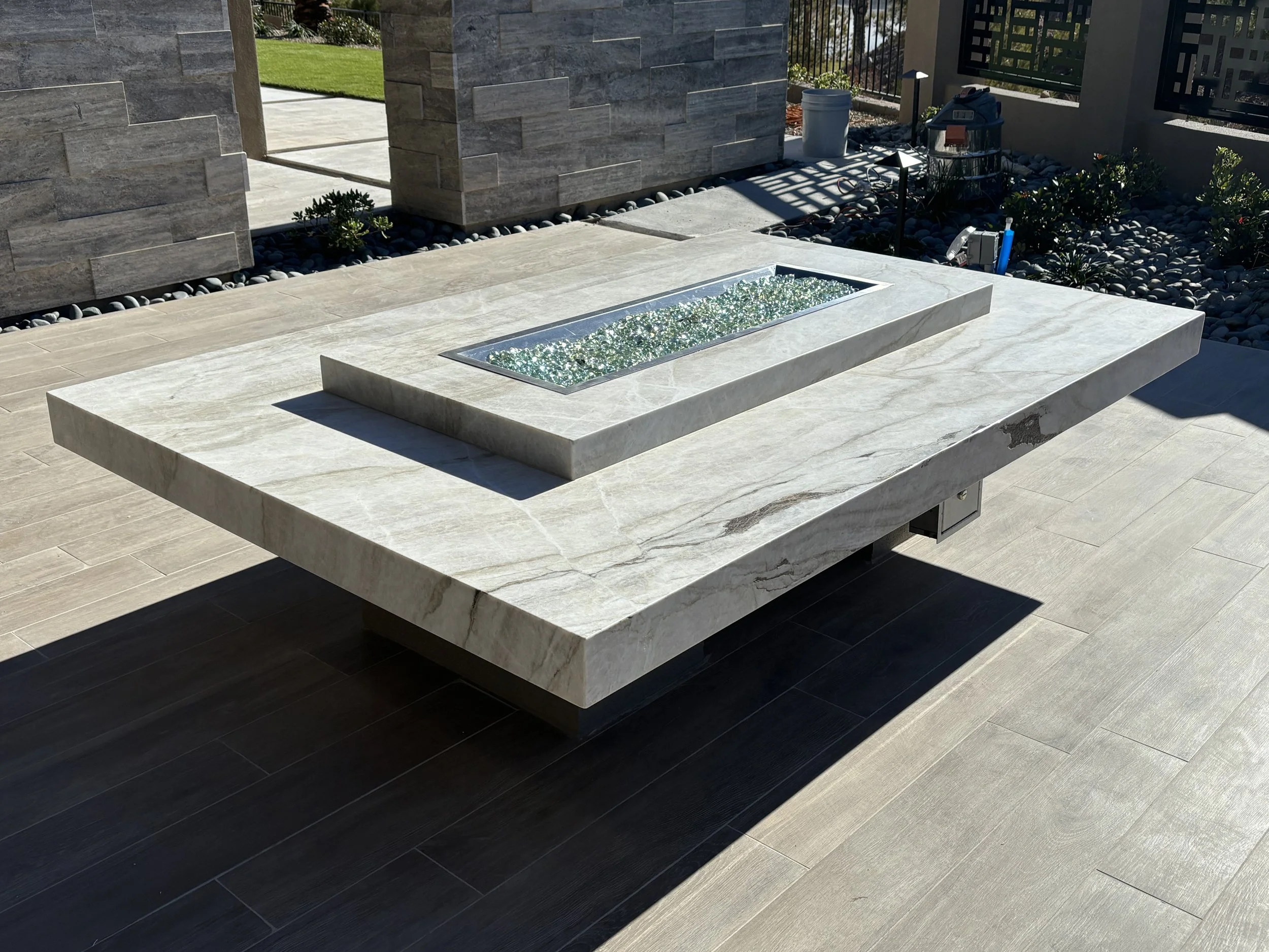 Modern outdoor rectangular stone fire pit table with glass pebbles, placed on a patio with stone tile flooring and landscaped garden area.