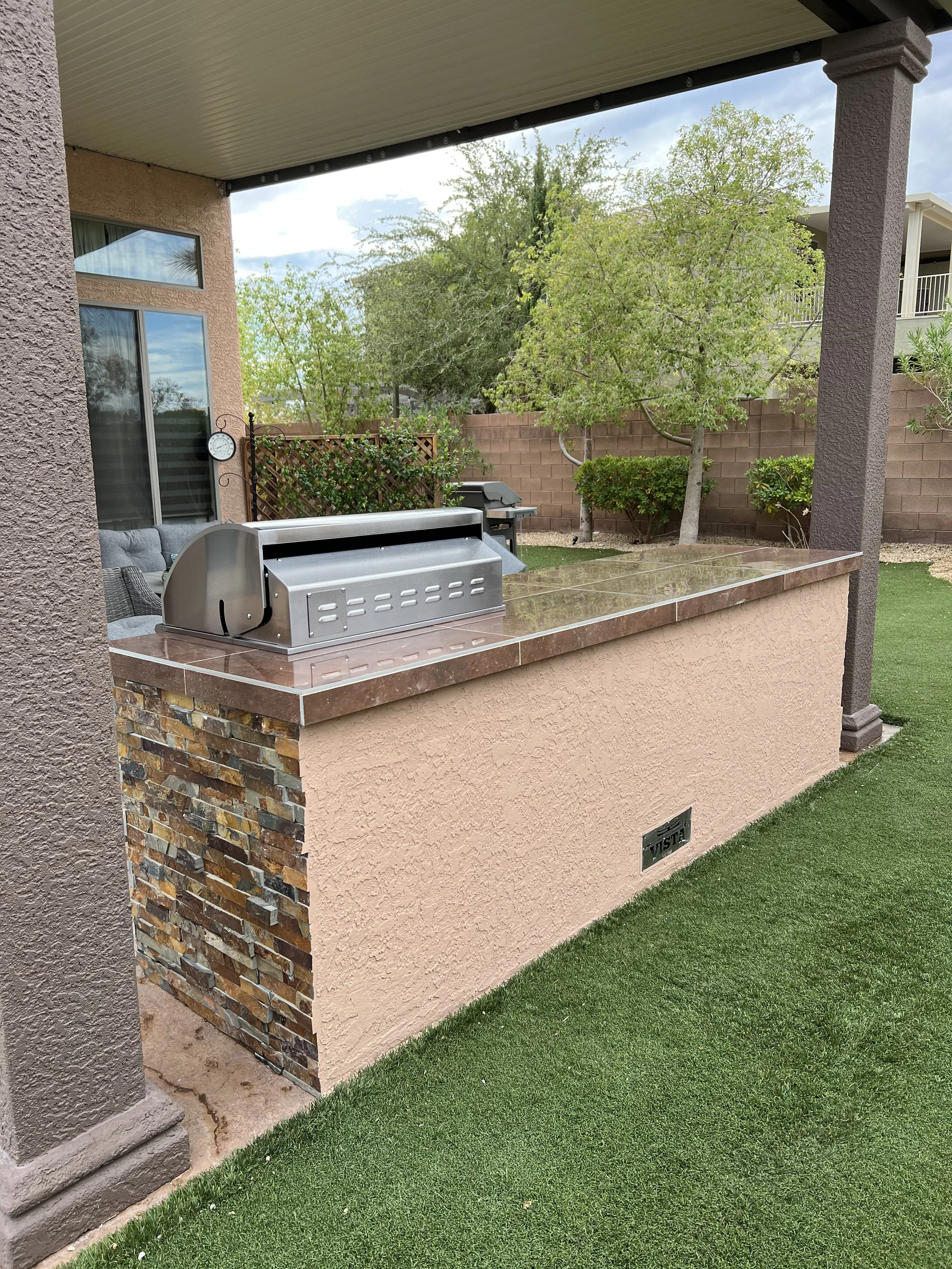 Outdoor patio with a built-in grill, featuring stone and stucco design, under a covered roof. There's artificial grass, trees, and a brick wall in the background.