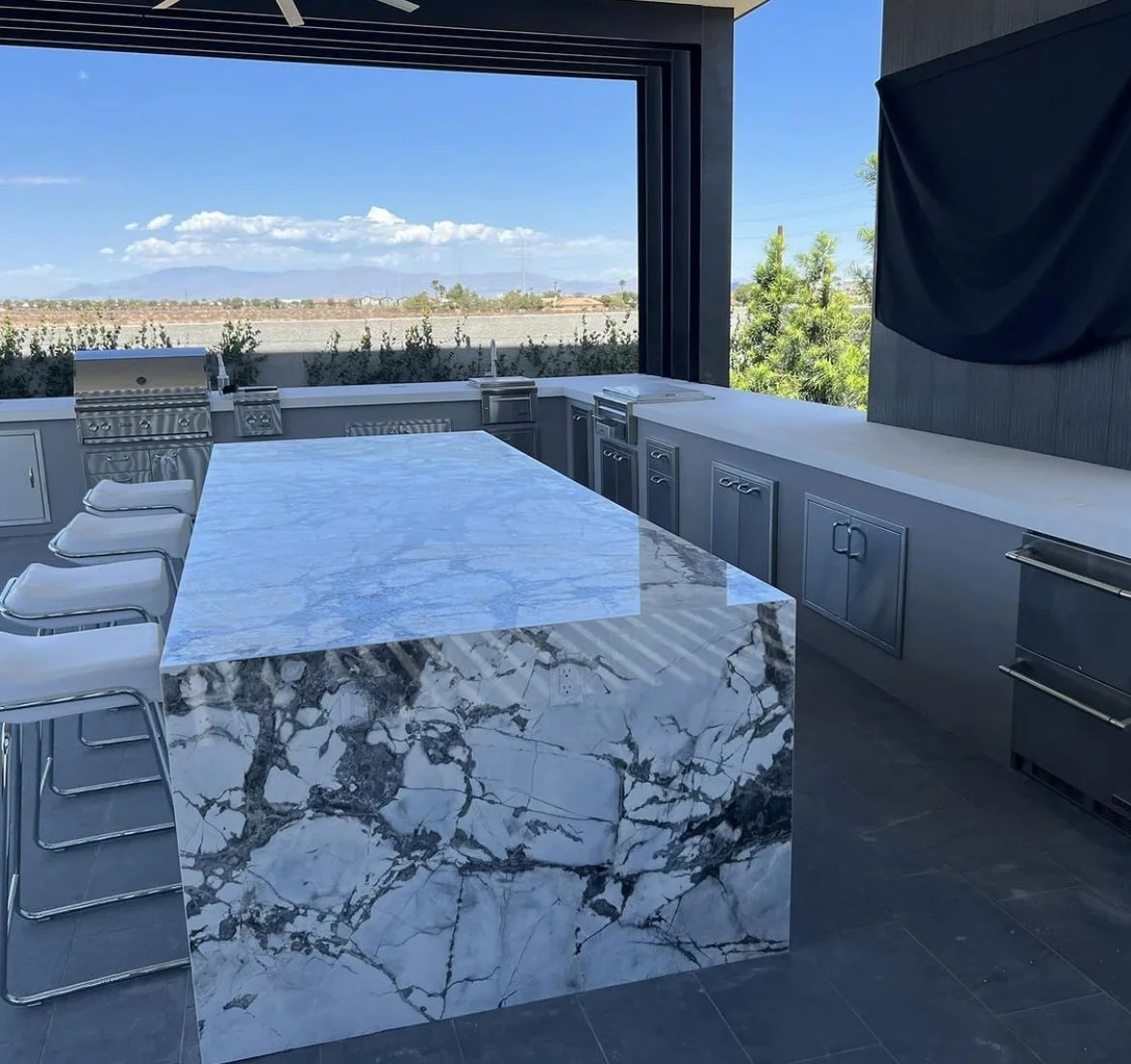 Modern outdoor kitchen with a large marble island, bar stools, stainless steel appliances, and a scenic landscape view.