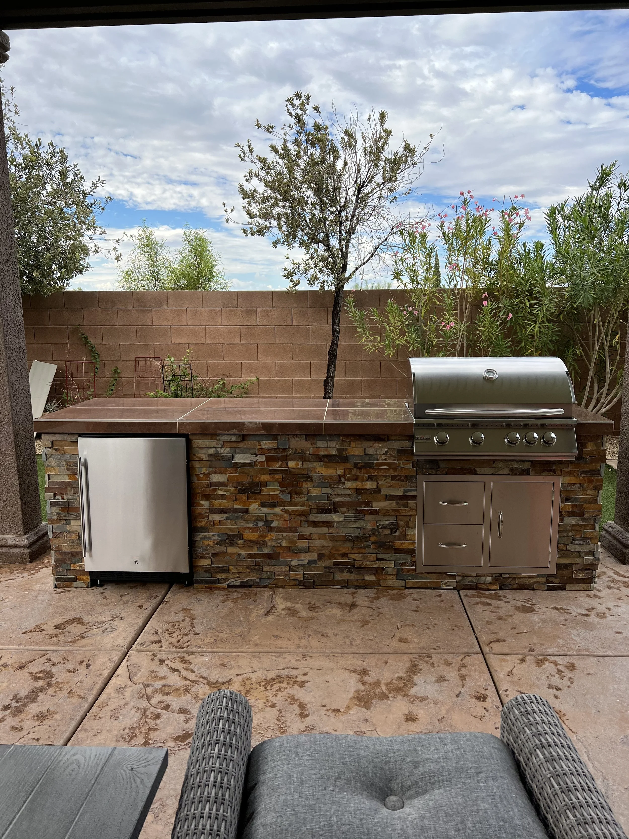 Outdoor patio with a built-in grill and refrigerator adjacent to a stone wall, surrounded by trees and plants under a cloudy sky.