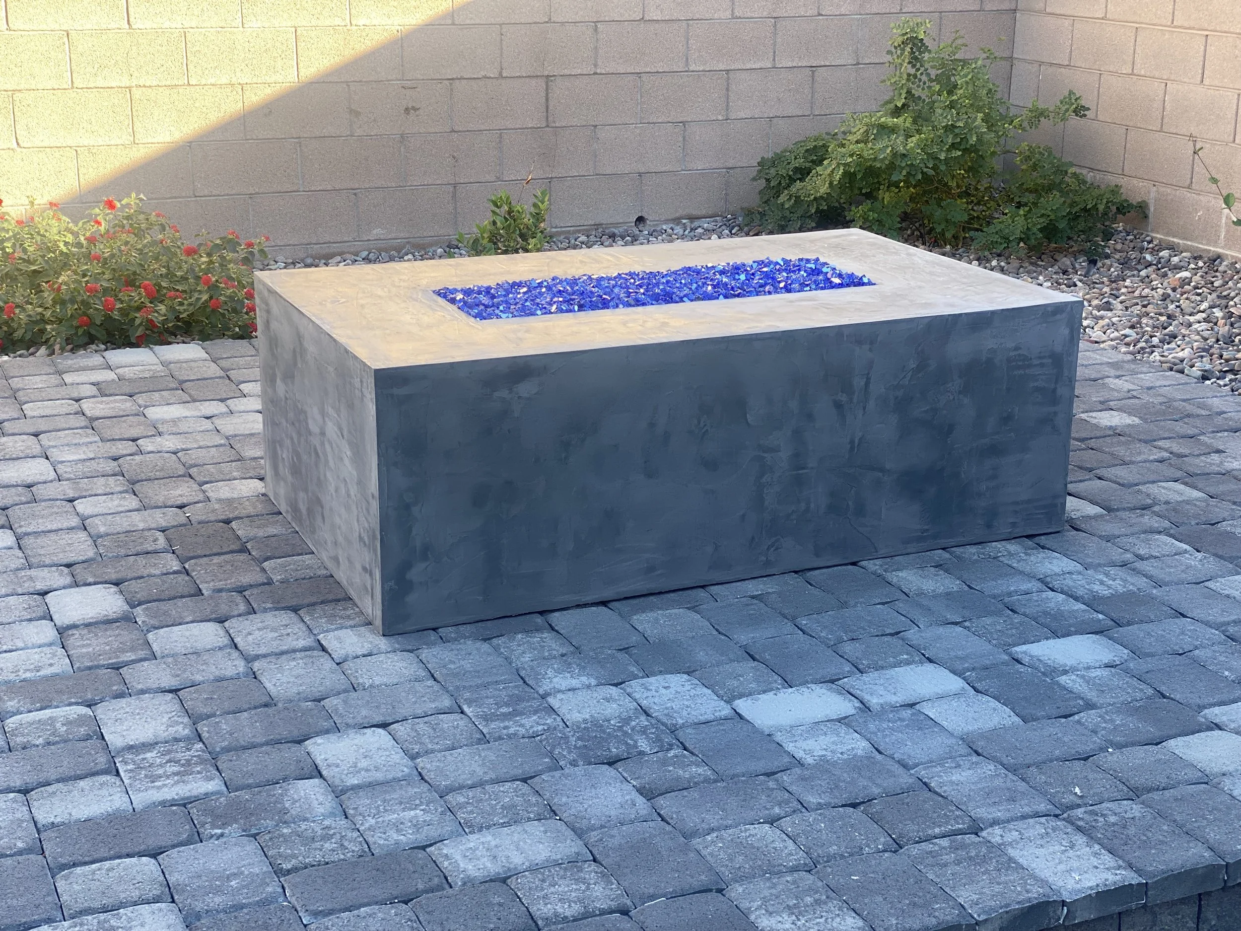 Rectangular concrete fire pit with blue glass stones on a paved patio.