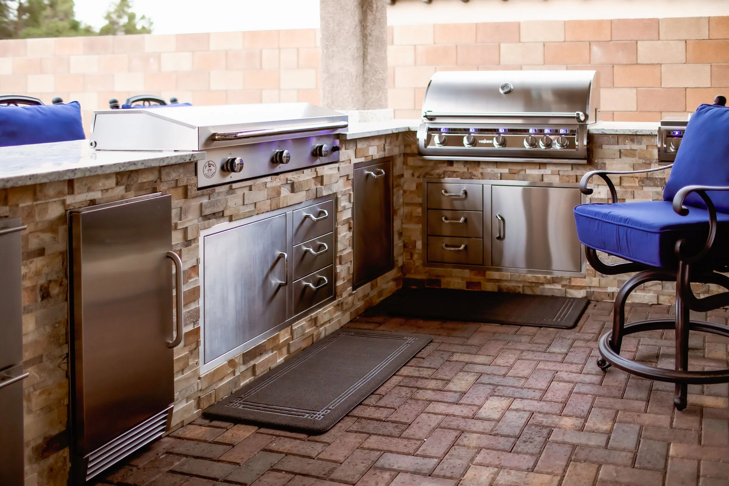 Outdoor kitchen with stainless steel appliances, including a grill and drawers, surrounded by stone brickwork; blue cushioned chair; brick flooring.