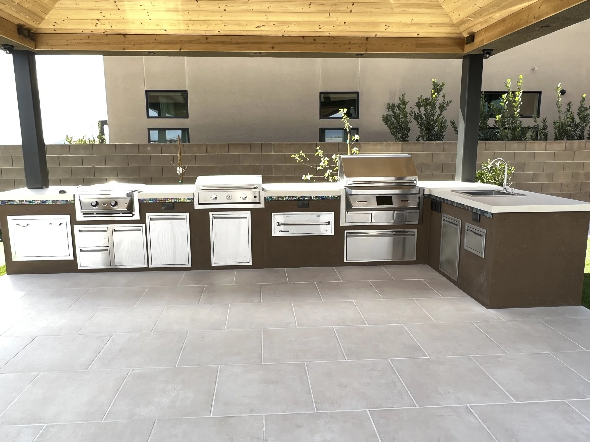 Outdoor kitchen with stainless steel grill, appliances, sink, and brown cabinetry under a wooden pergola.