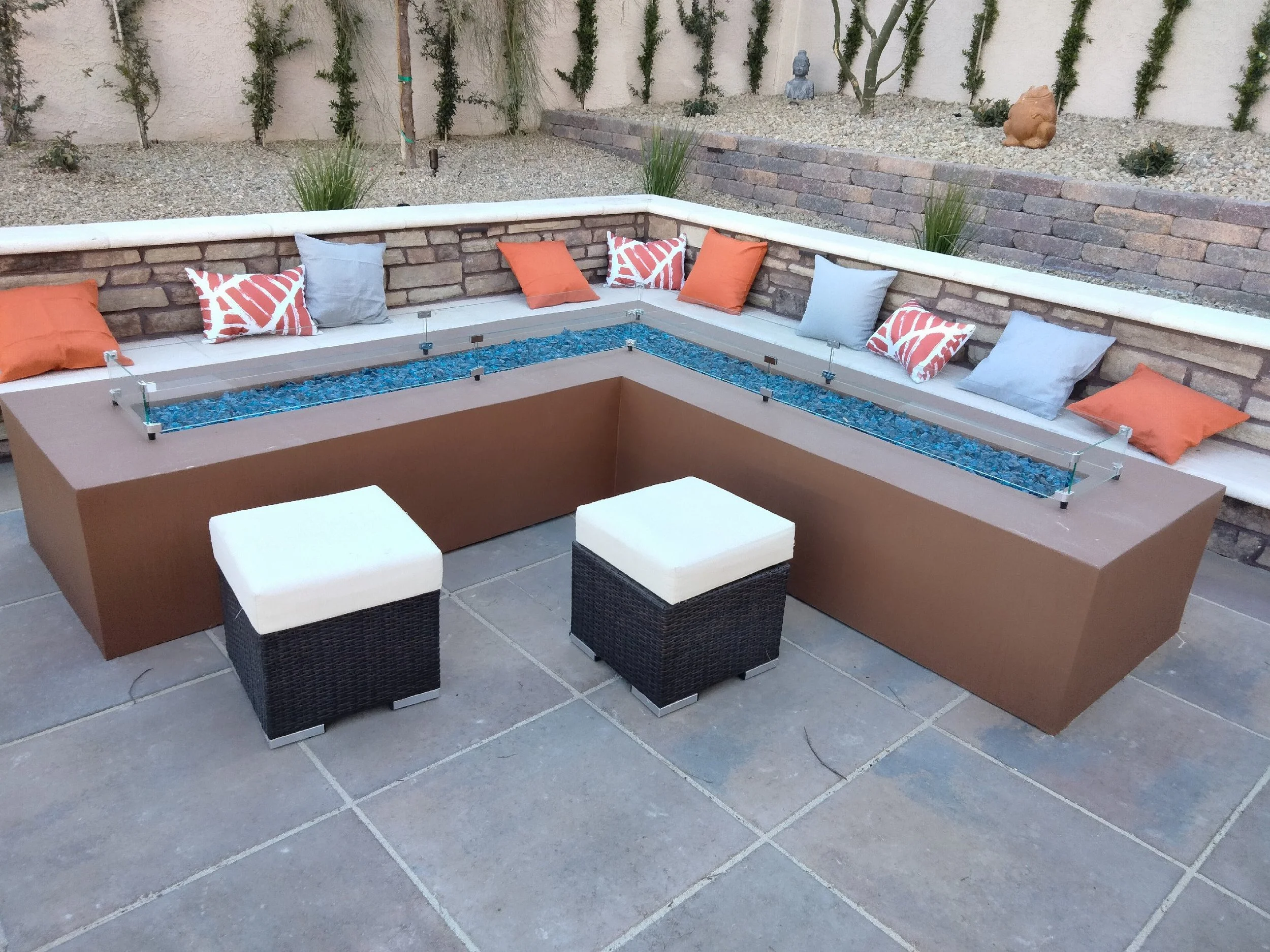Outdoor patio with a brown fire pit table, blue glass pebbles, surrounding bench seating with orange and gray pillows, and two white cushioned cube seats on stone tile floor.