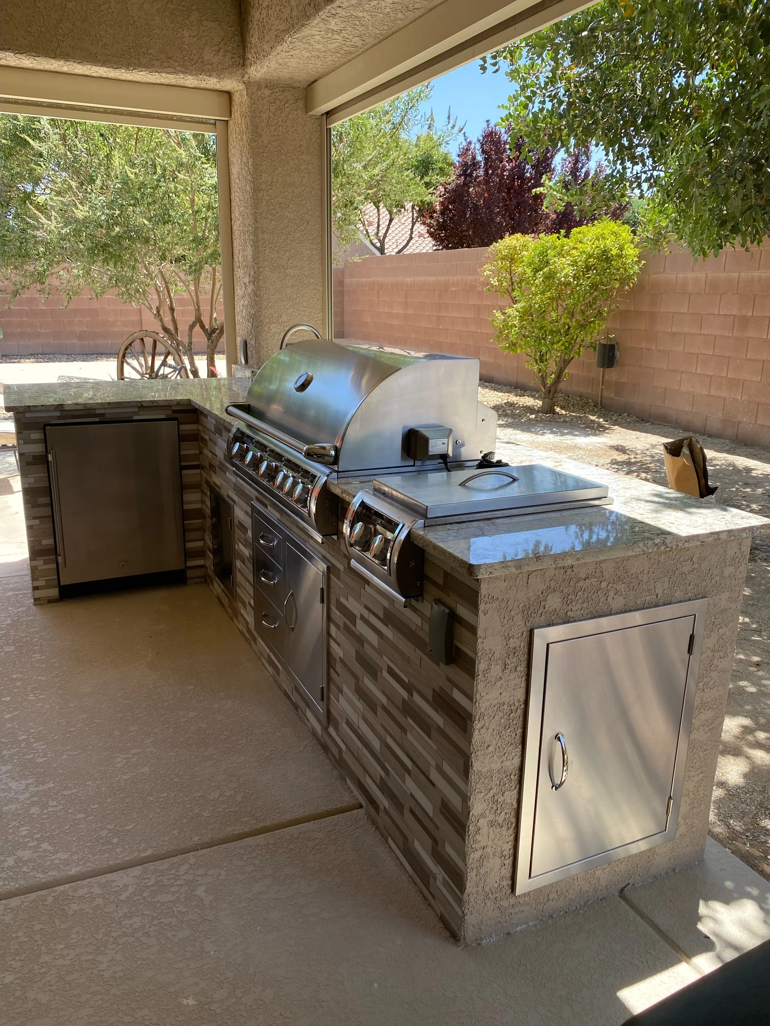 Outdoor kitchen with stainless steel grill, countertop, and cabinets in a backyard setting with trees and a stone wall.
