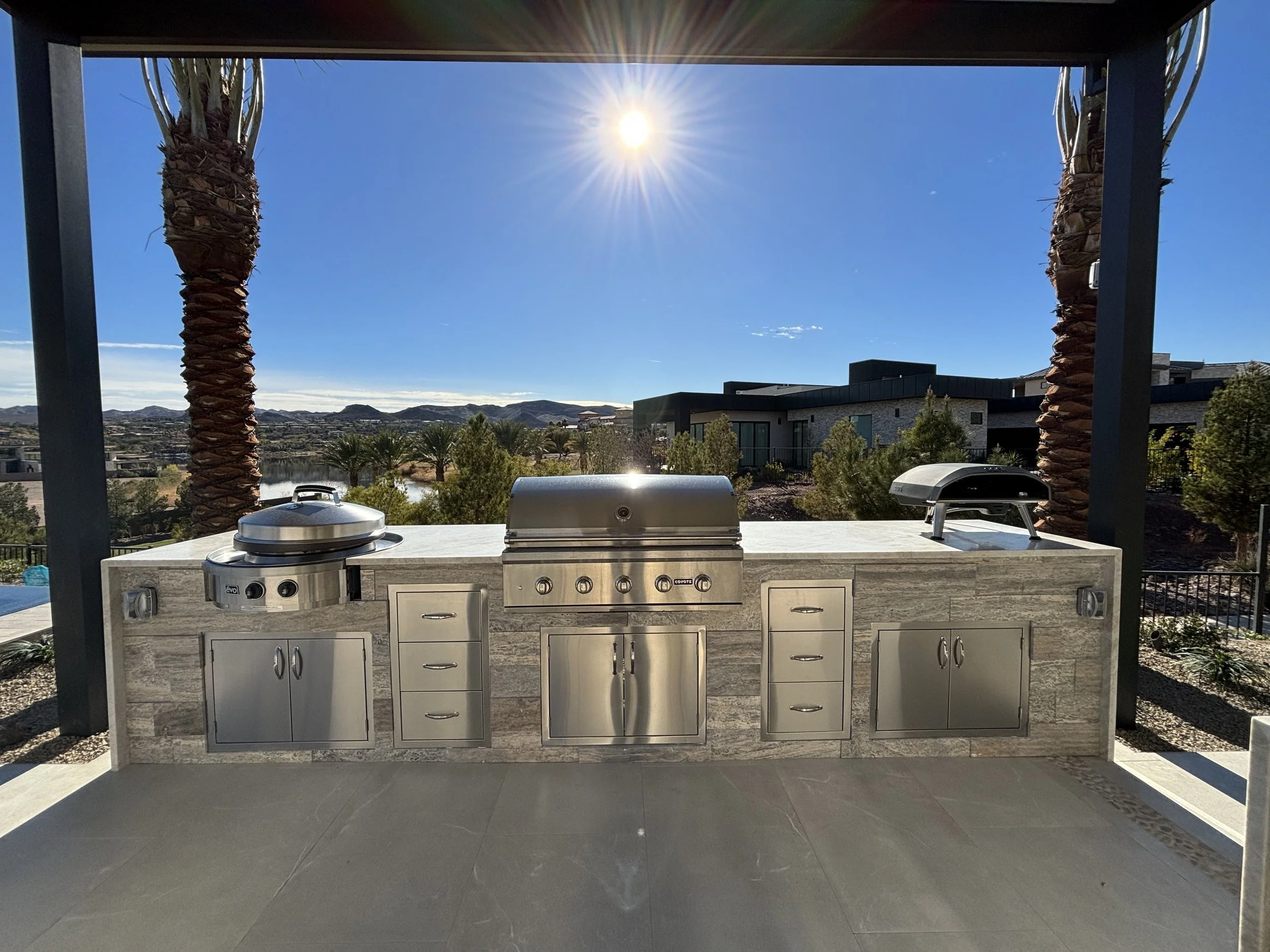 Outdoor kitchen with stainless steel appliances, grill, and storage cabinets, surrounded by a landscape featuring palm trees and houses, under a clear blue sky with the sun shining.